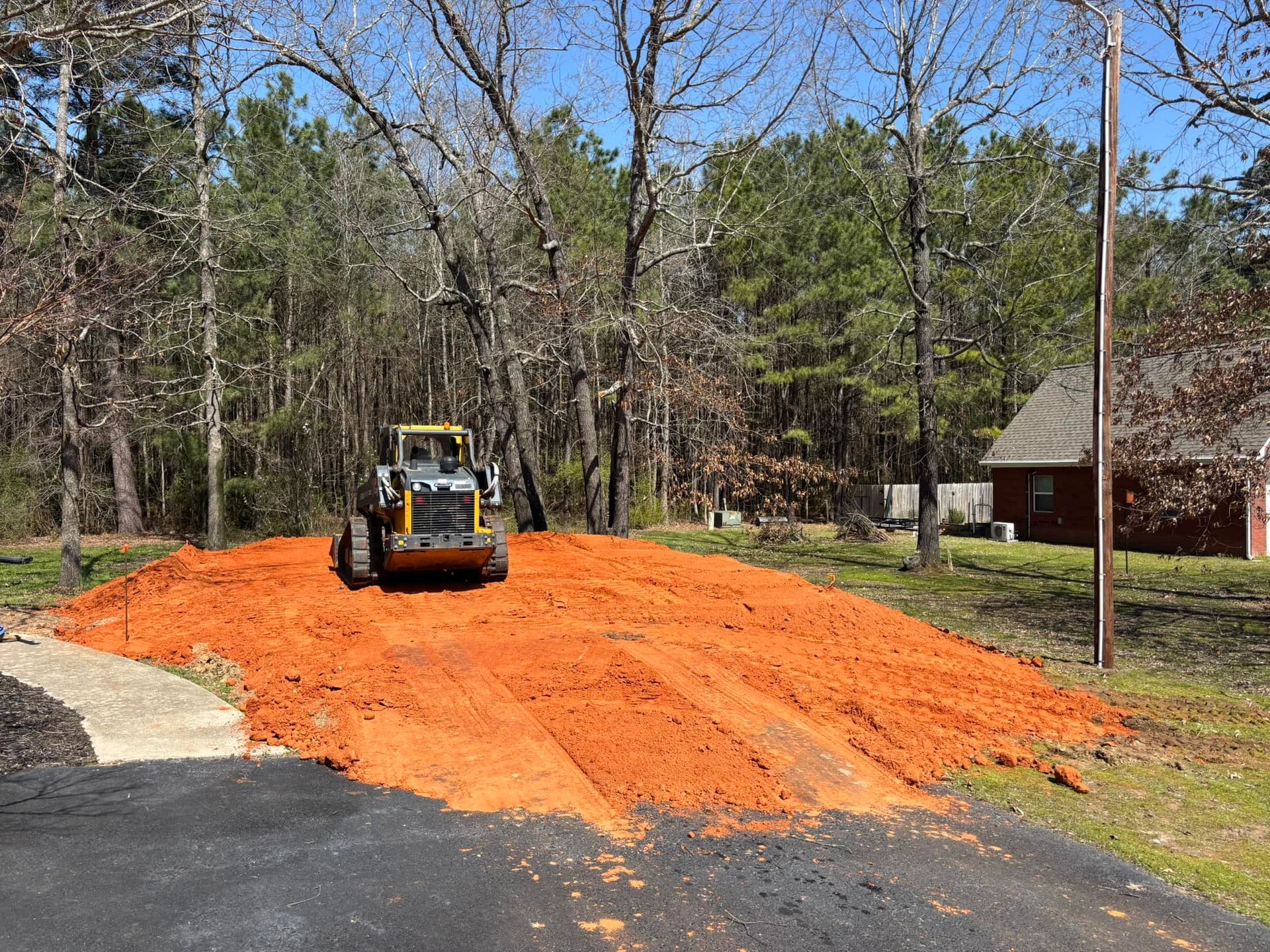 Yellow construction vehicle compacting red soil on a driveway, with trees and a house in the background.