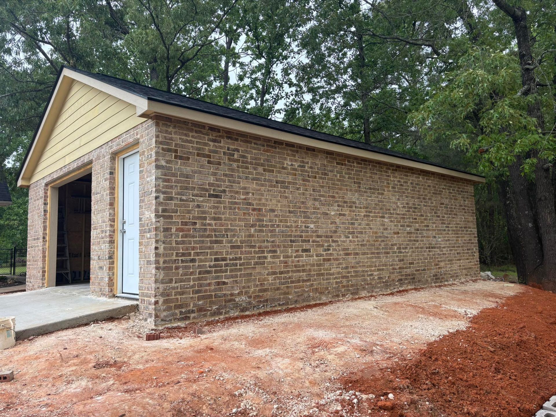 Brick building with two garage bays, a door, and a concrete approach. Surrounded by dirt and trees.