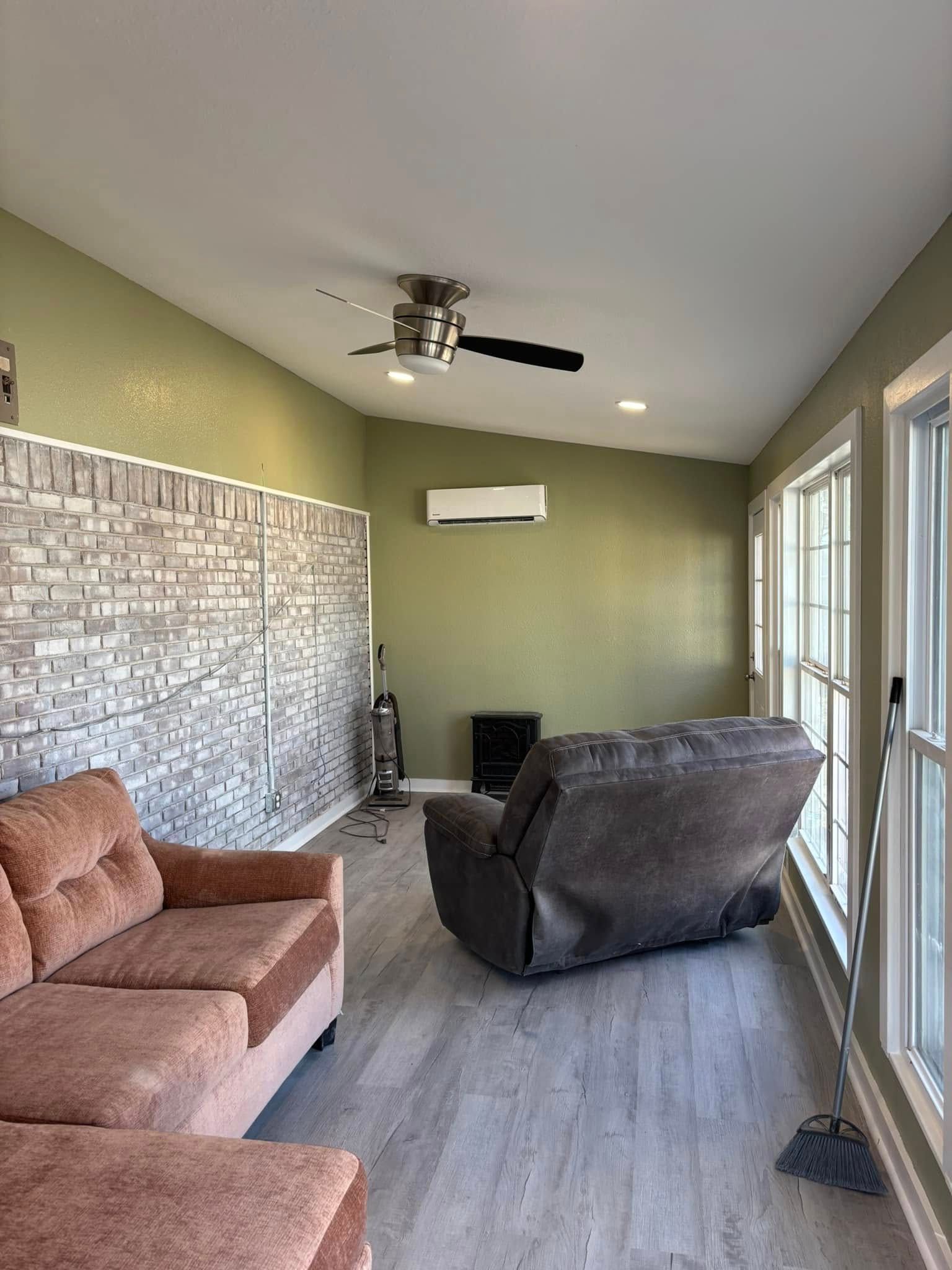 A room with a sofa, recliner, and windows. Light gray flooring, olive green walls, and a brick wall accent.