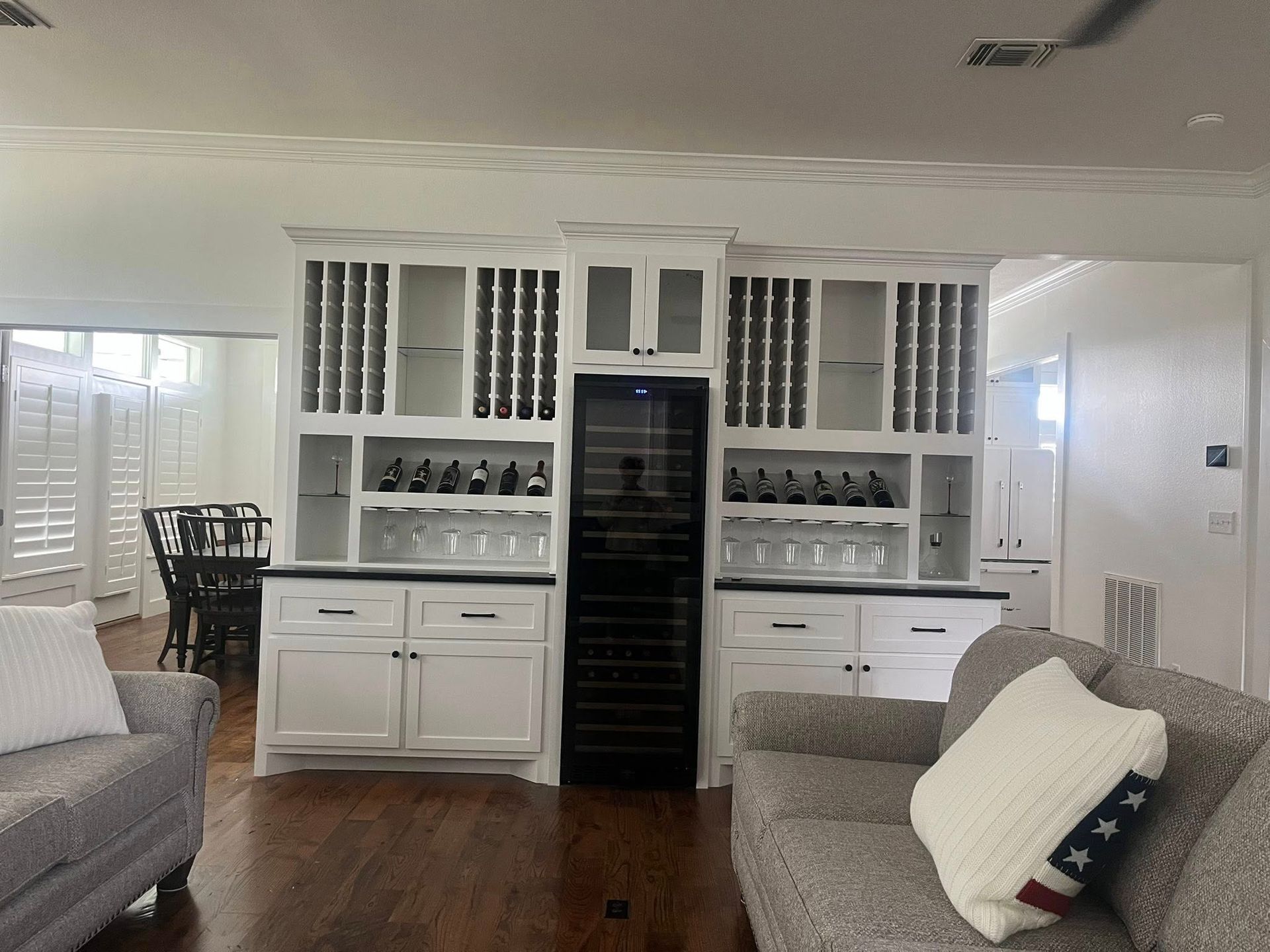 White built-in cabinet with wine storage and cooler. Seating area with sofa and chairs. Dark wood flooring.