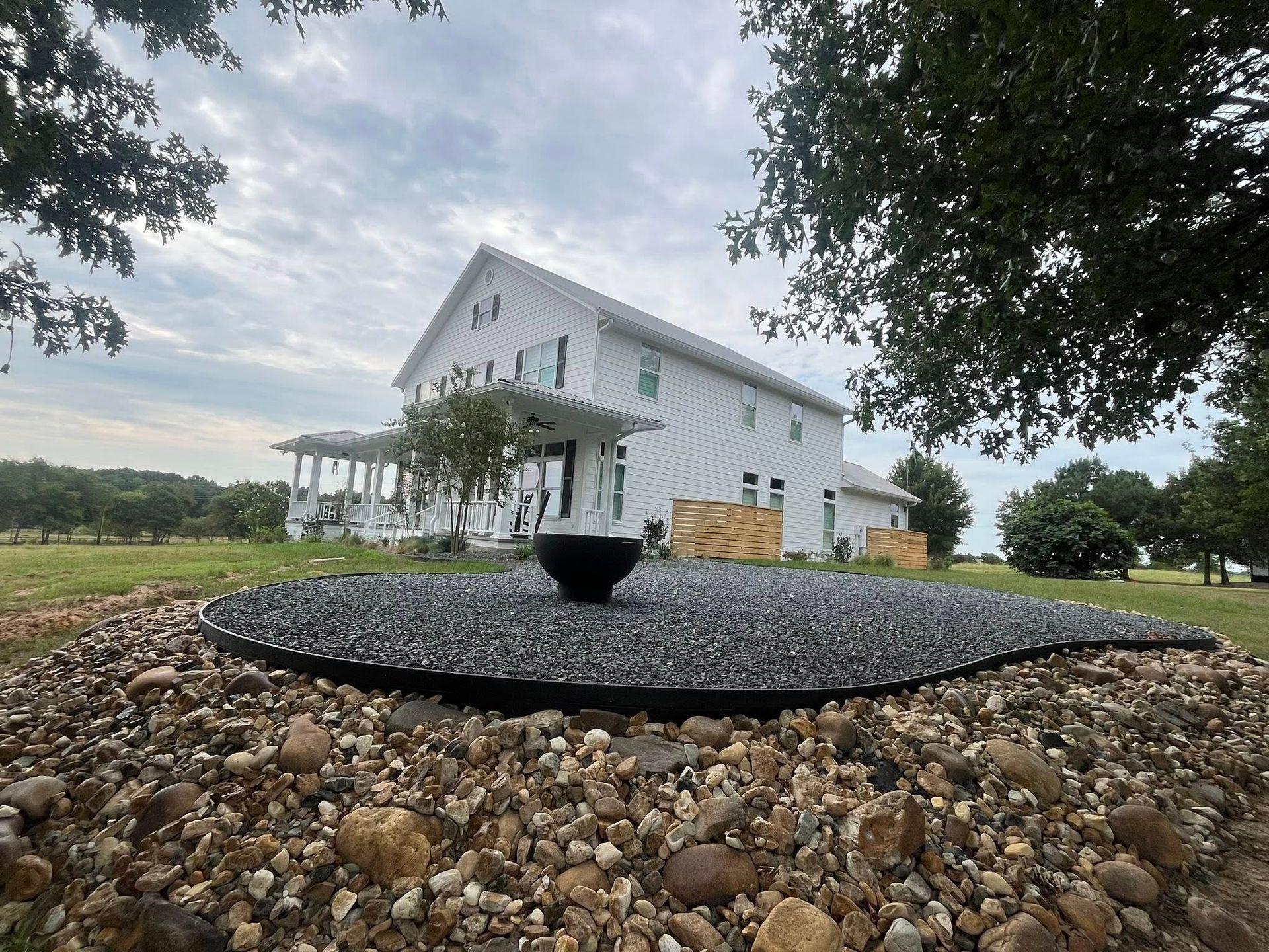 White farmhouse with a black, teardrop-shaped water feature in front. Gravel landscaping and cloudy sky.