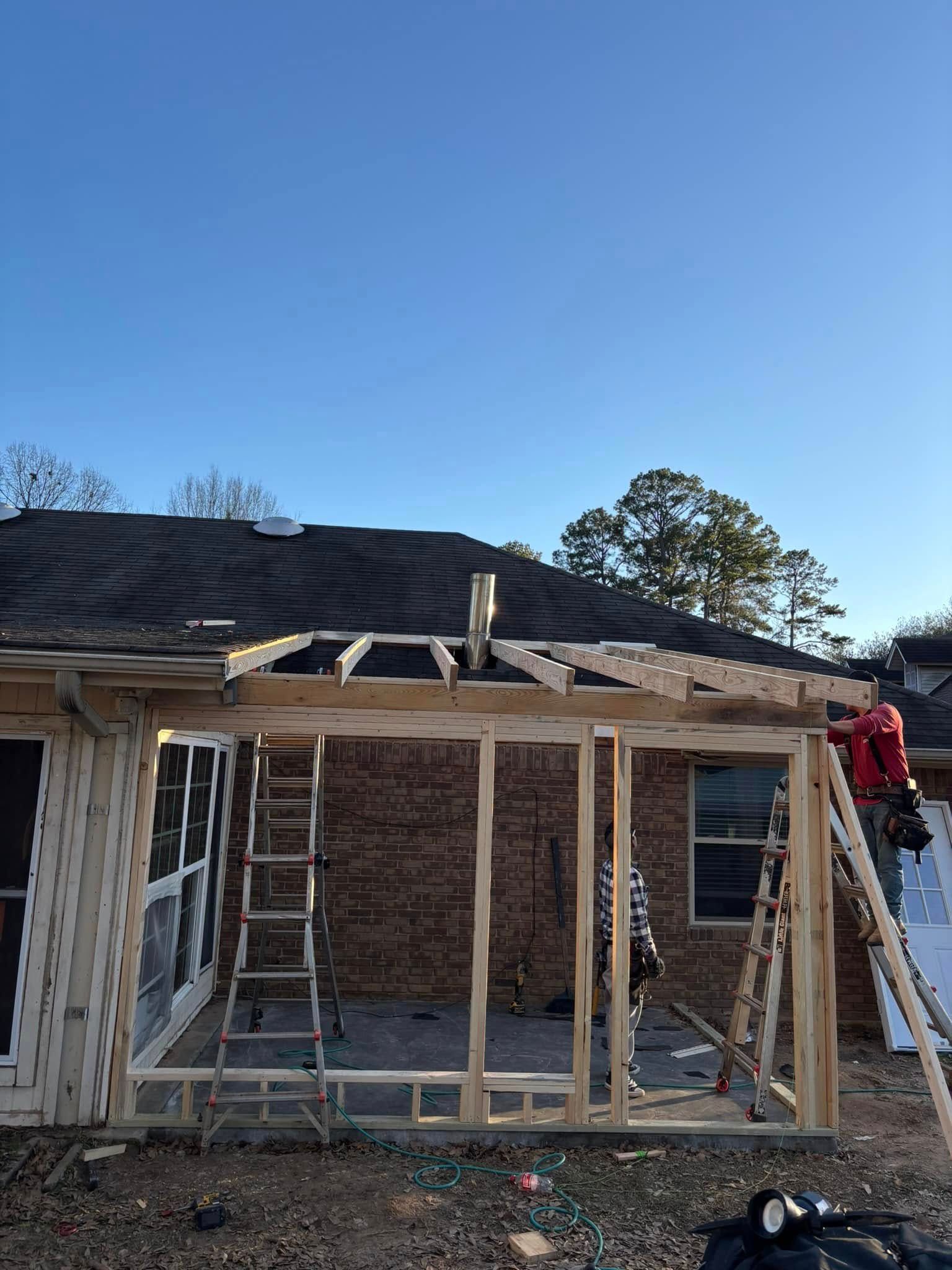 Construction of a wooden framed sunroom addition to a brick house. A person stands on a ladder.