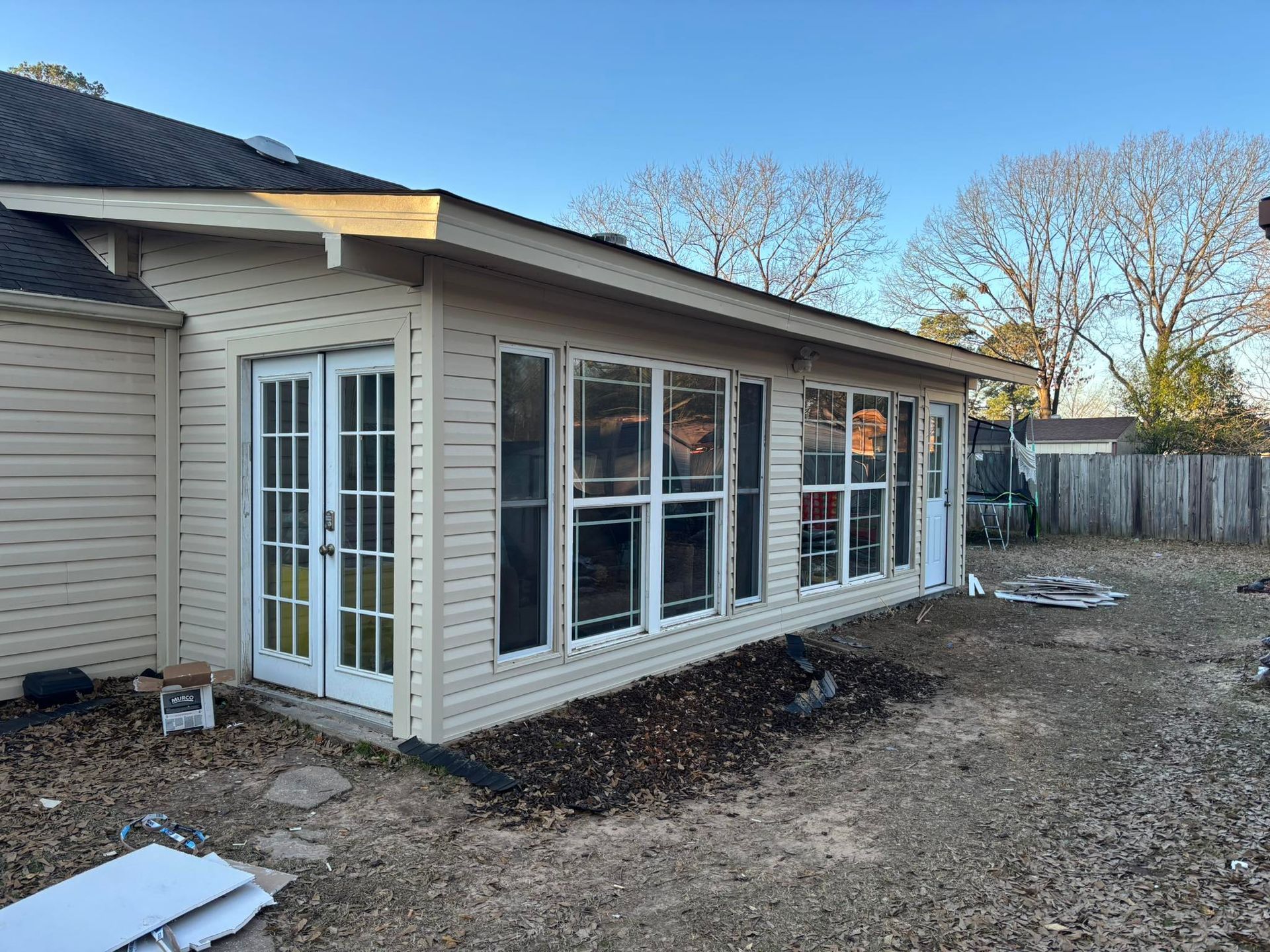 Exterior of a sunroom with French doors and large windows, attached to a house with beige siding. Back yard visible.