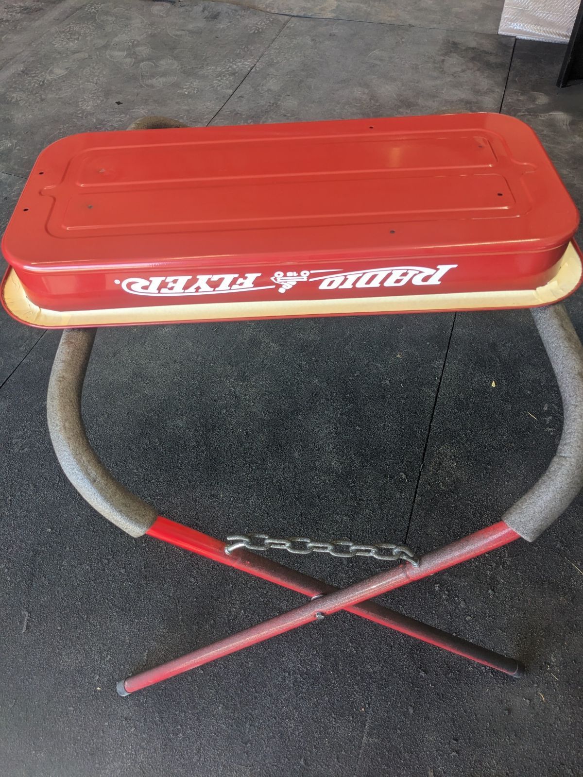 Red Radio Flyer wagon body converted into a seat, supported by metal legs; gray padding on the legs, with a chain.