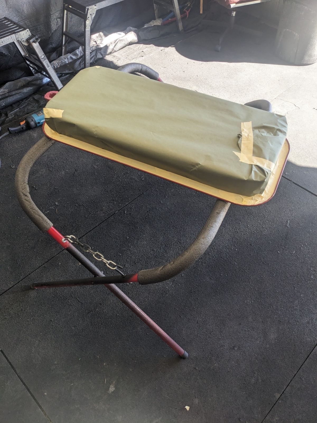 A makeshift weightlifting pad on a thick curved pipe, with two red-tipped sticks lying nearby on a black gym floor.