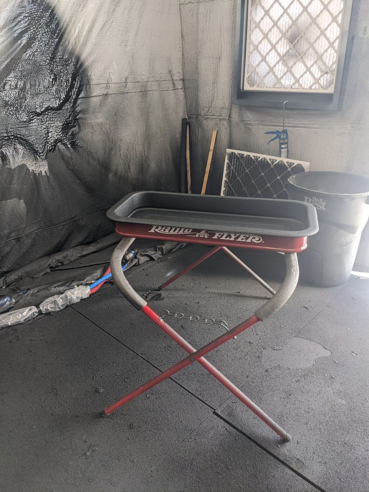 Red wagon tray on a red folding stand in a dark room; window, bucket, and tools in the background.