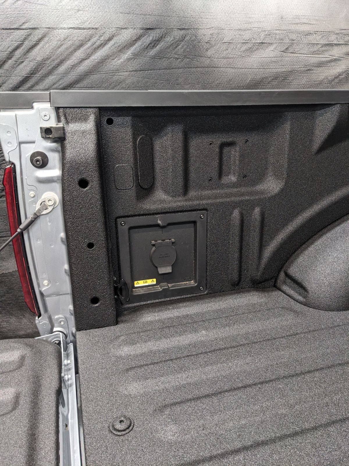 Truck bed with a black 120V power outlet in the wall. Silver truck body and black bed liner.