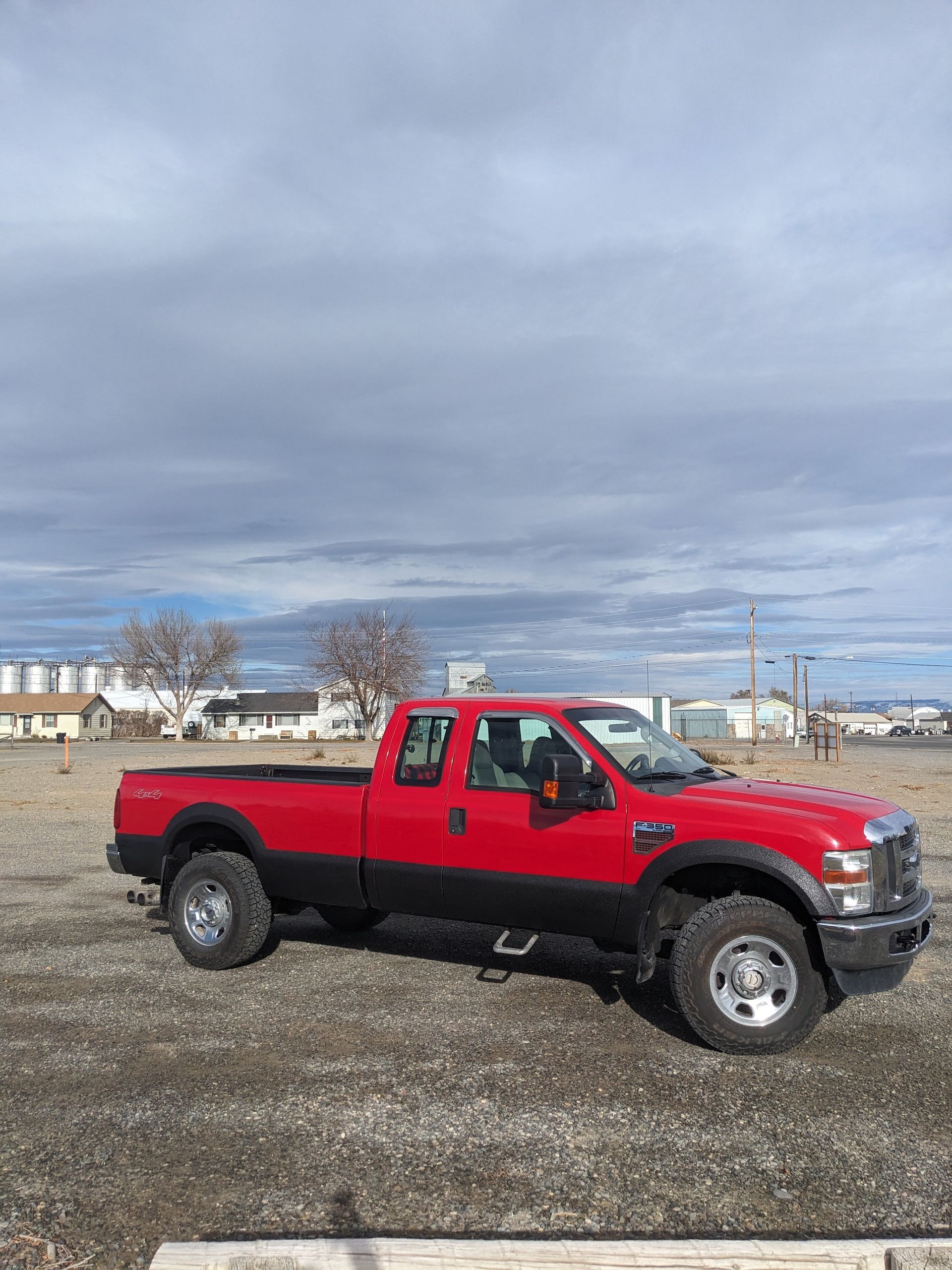 Red and black Ford pickup truck parked in a gravel lot under a cloudy sky.