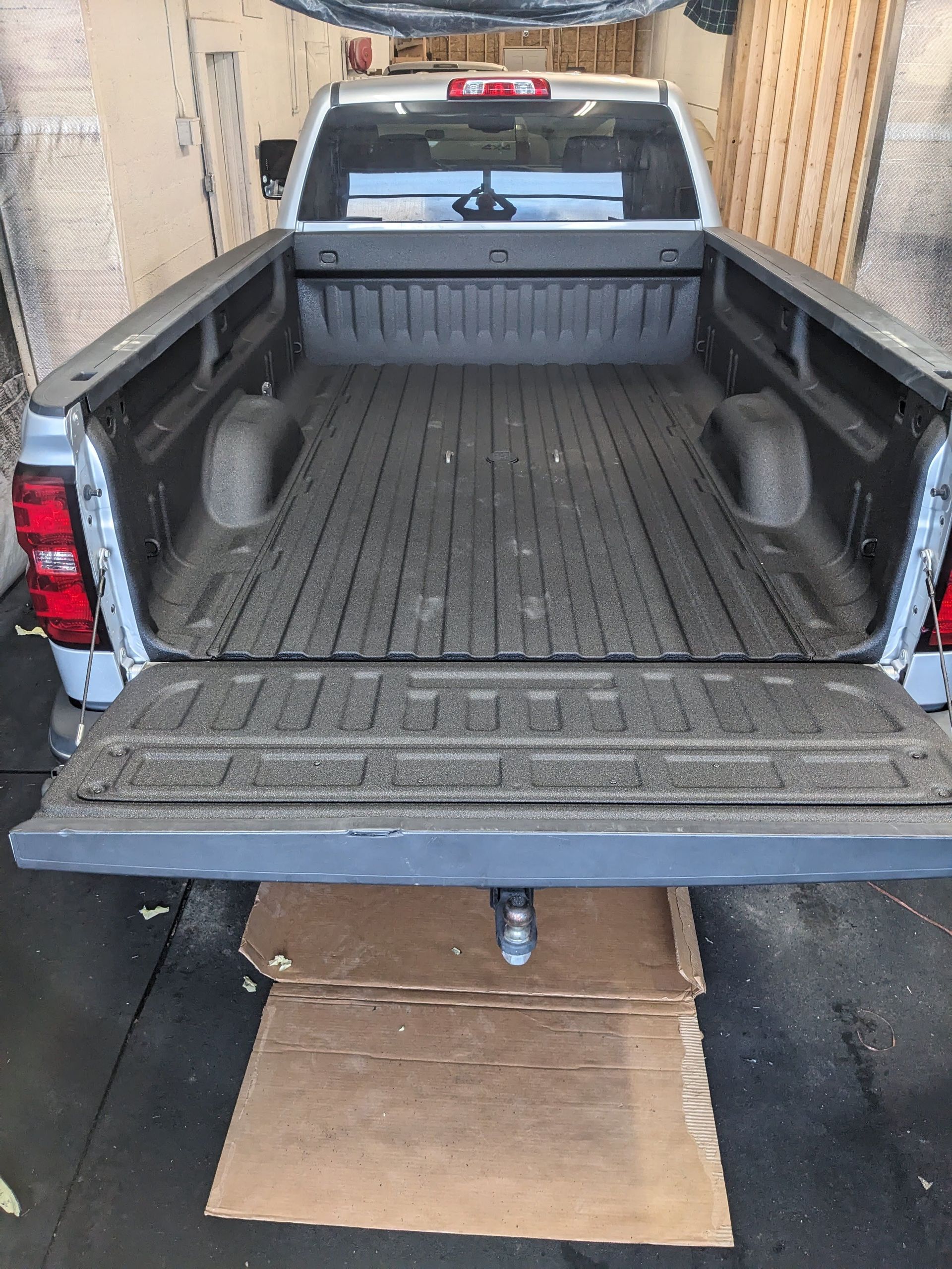Silver pickup truck bed, tailgate down. Cardboard covers the trailer hitch. Black bed liner.