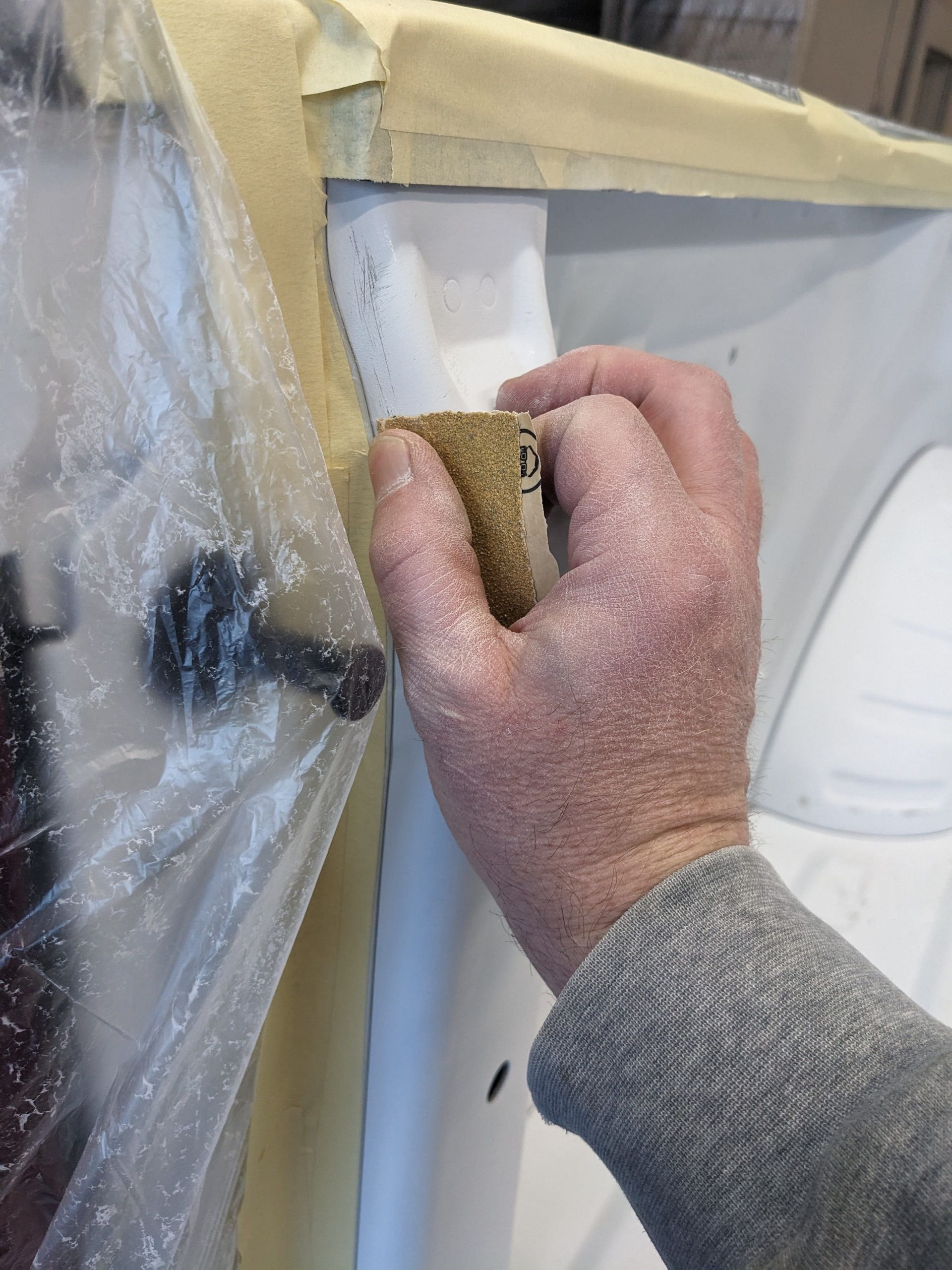 Person sanding a car body panel, inside a garage, wearing a grey sweater.