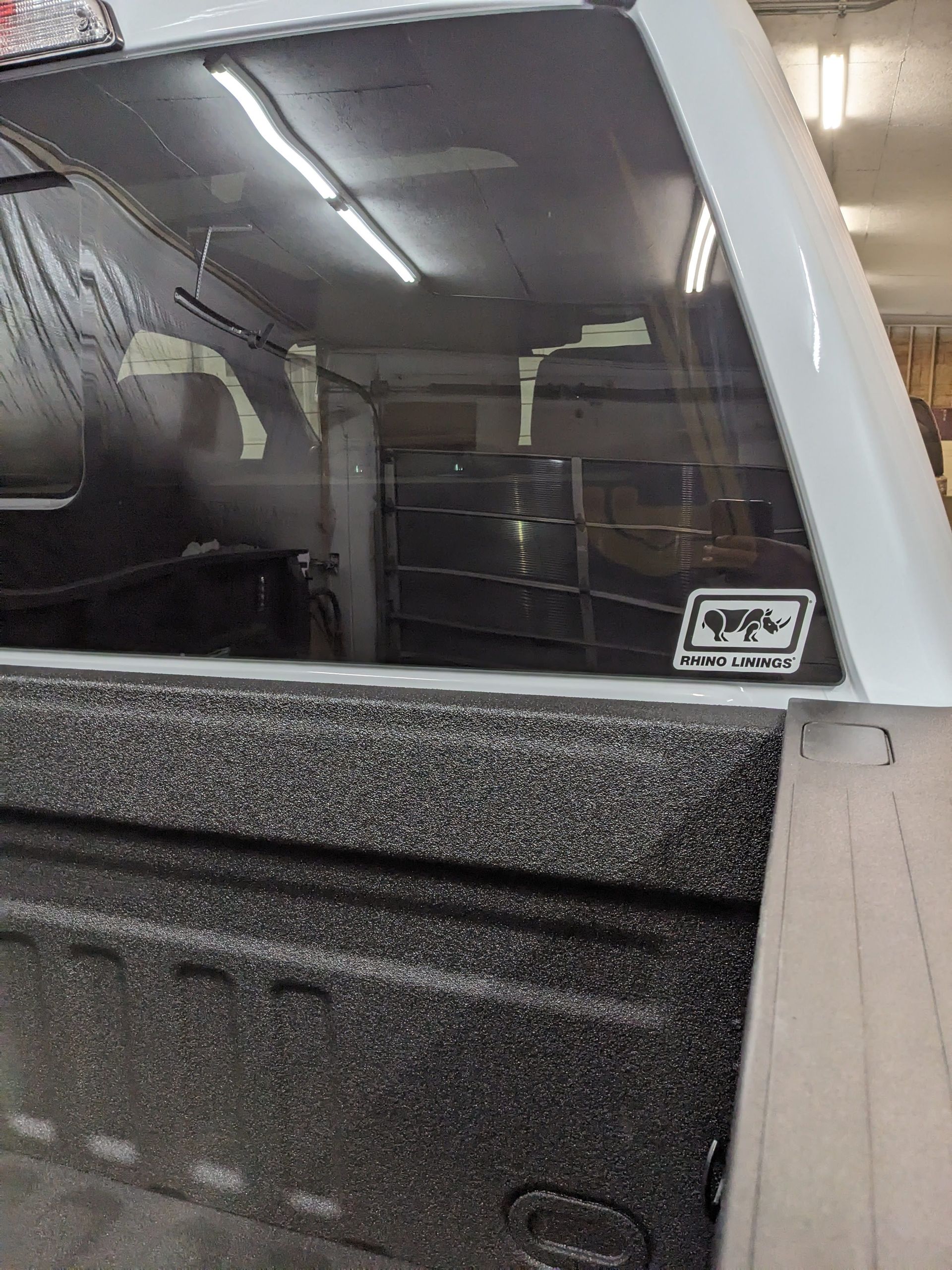 White pickup truck bed with black textured liner, rear window, and a sticker.