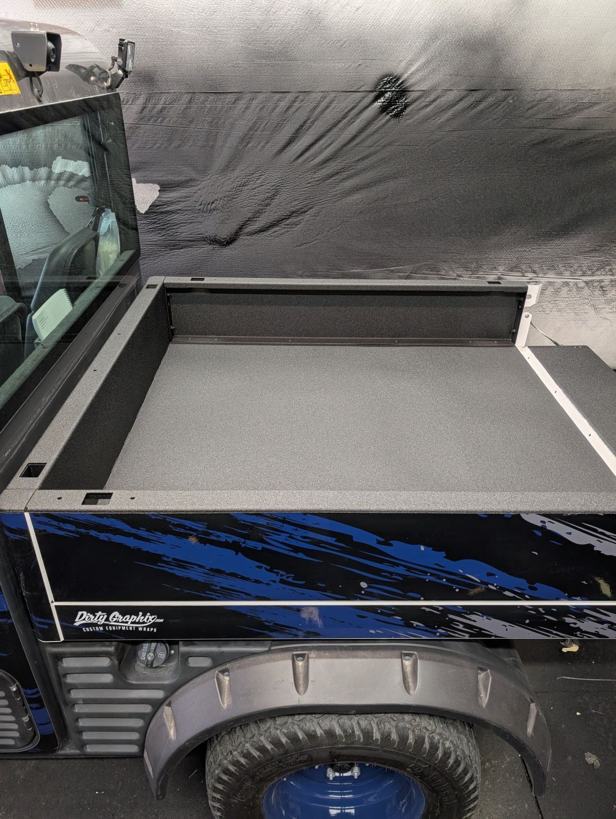Truck bed with a black bedliner, visible side panel with blue design, black tire.