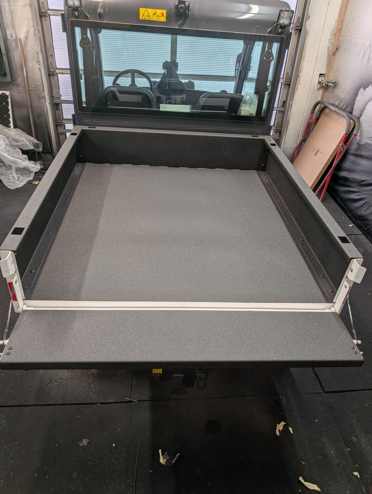 Empty, gray-carpeted utility vehicle bed, with open rear gate, against a background of the cabin.