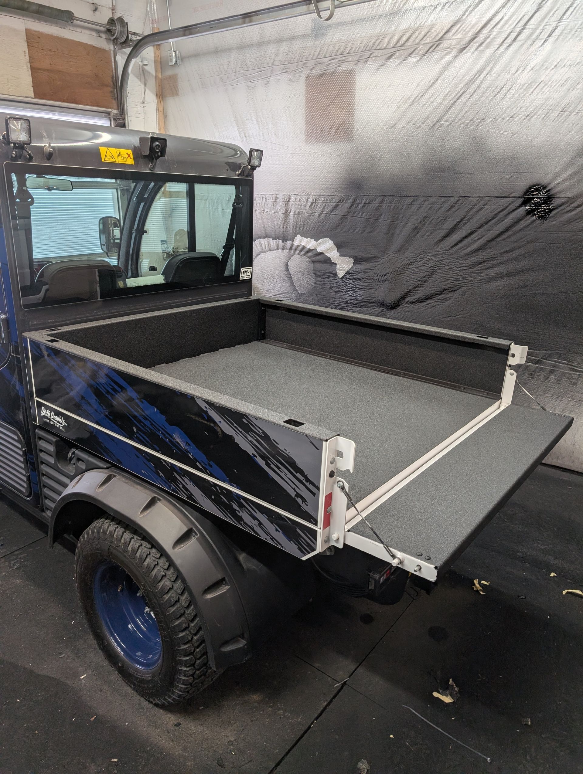 Blue utility vehicle with open bed, parked indoors. Bed has a textured surface. Black and gray color scheme.