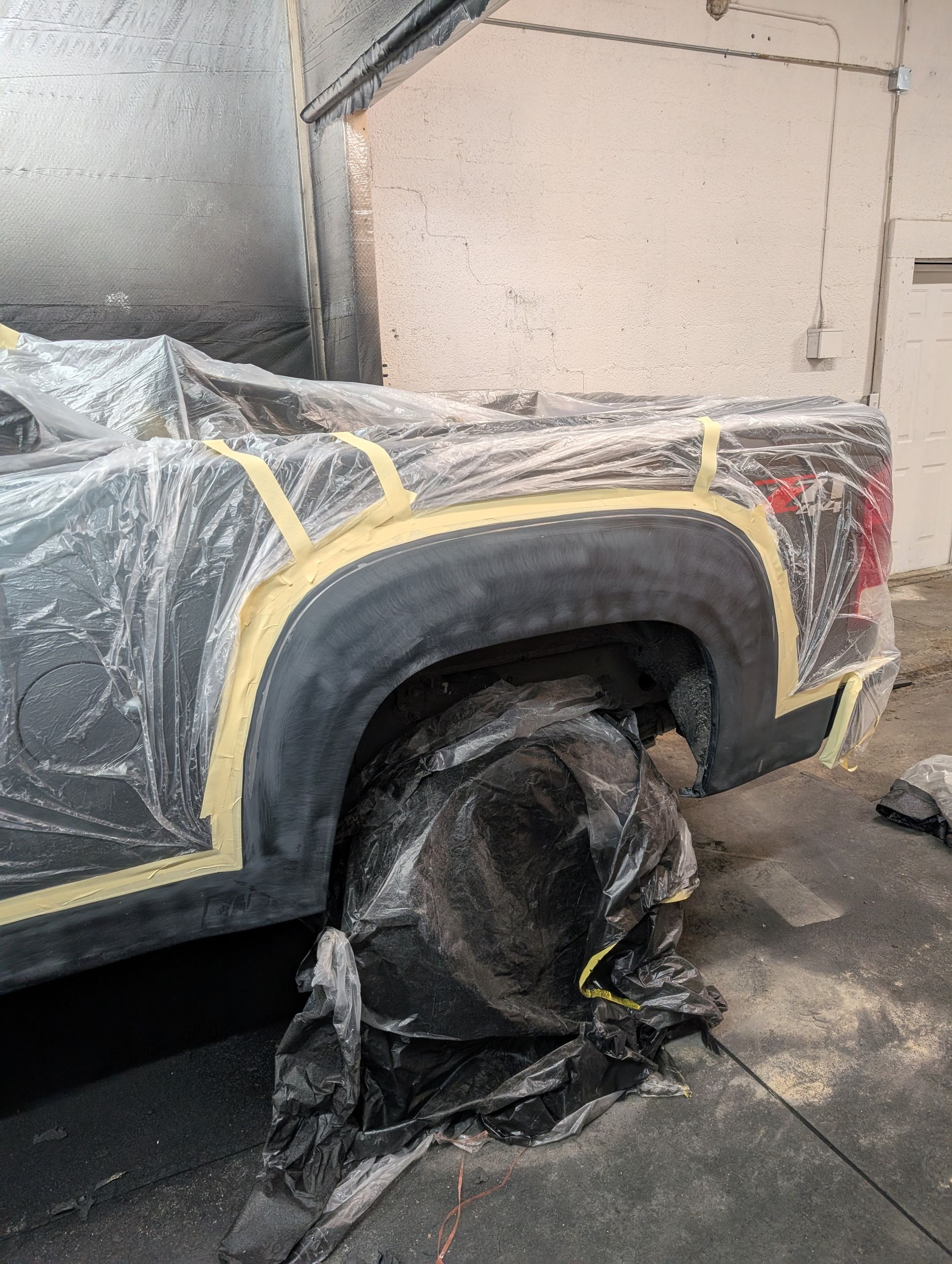 Truck bed prepped for paint; black fender, wheel covered, masked with yellow tape, inside a garage.