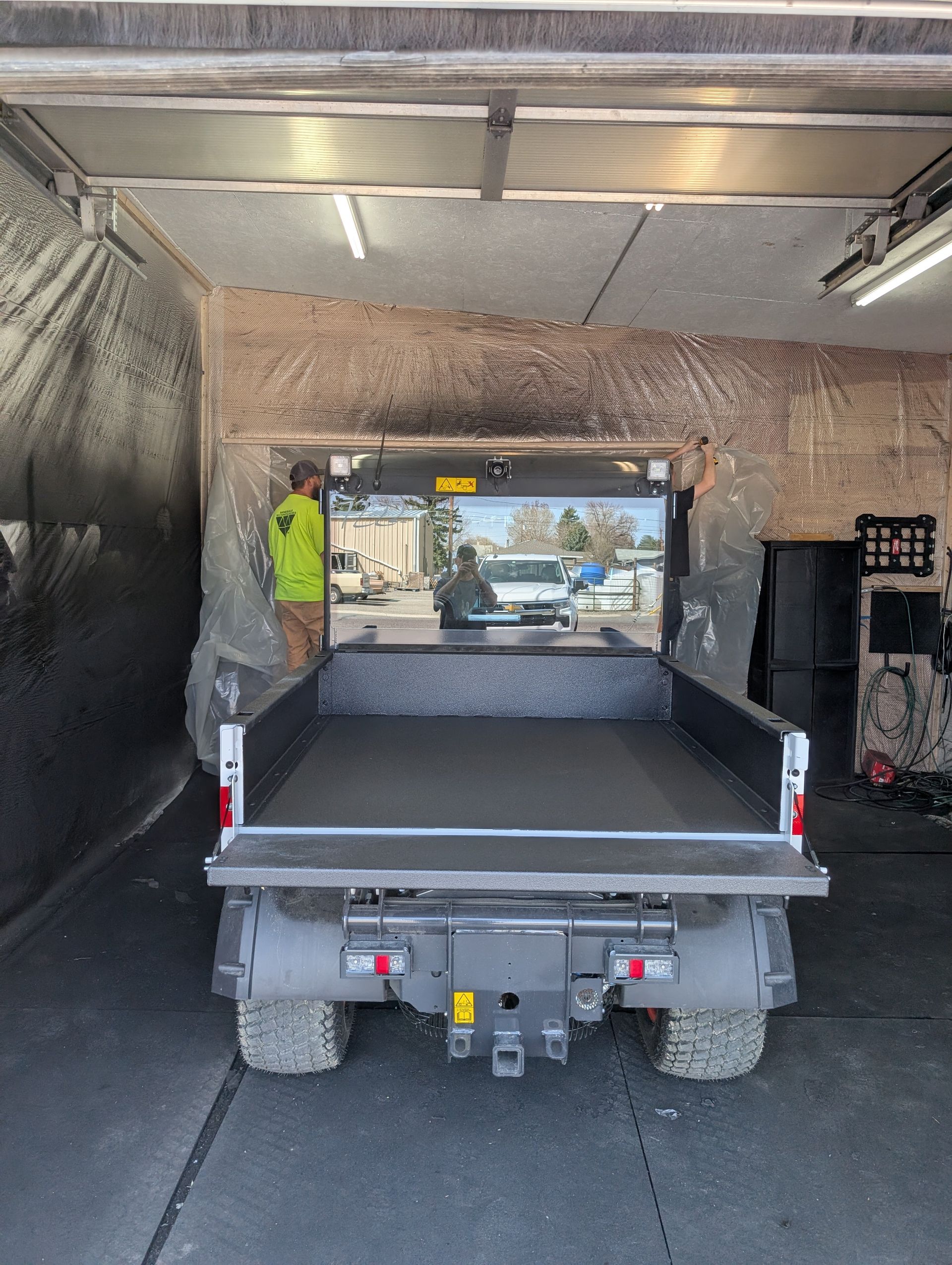 Utility truck with empty bed backed into garage. Worker standing near rear, another in cab.