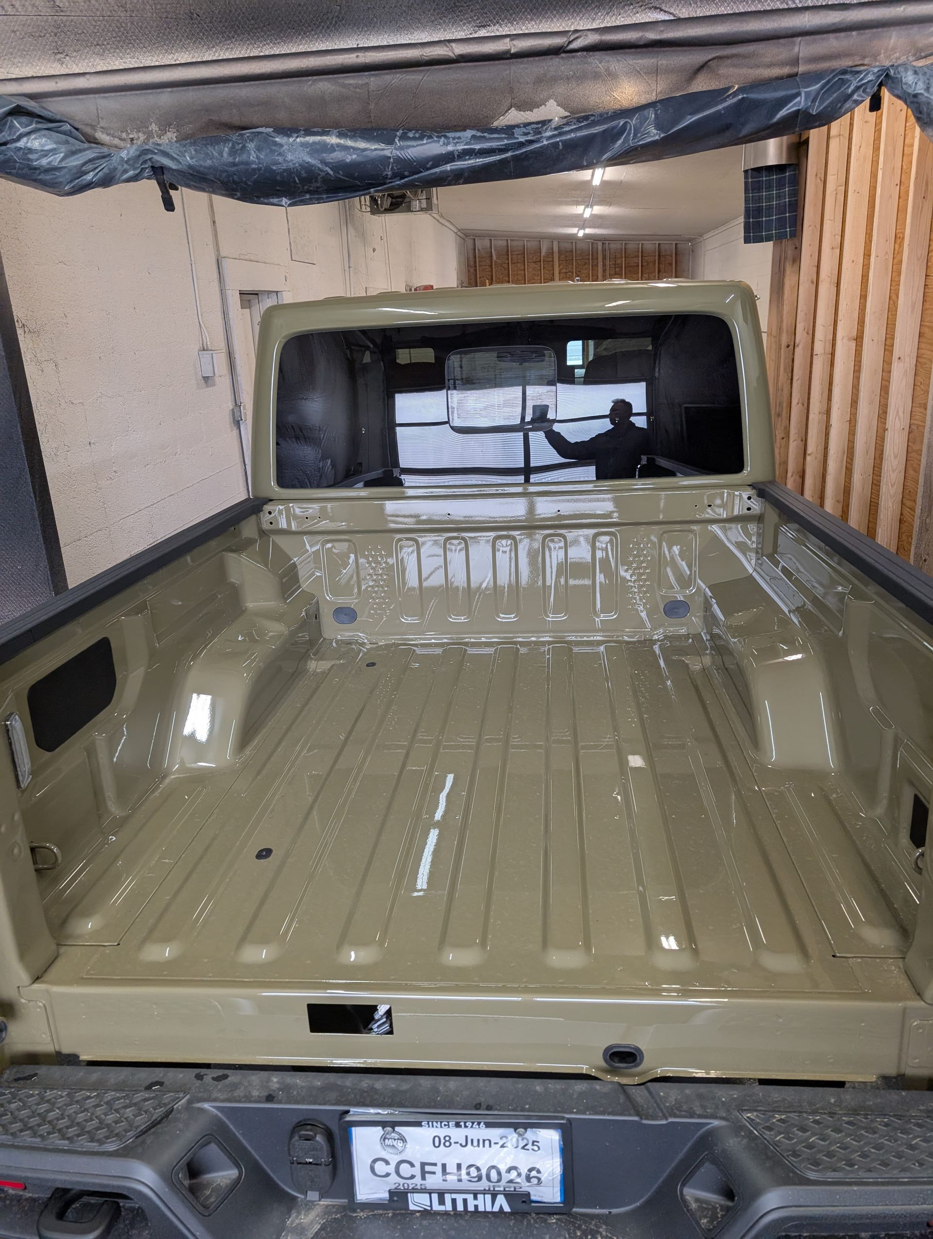 Olive green truck bed, rear view, inside a workshop. A person sprays a coating.