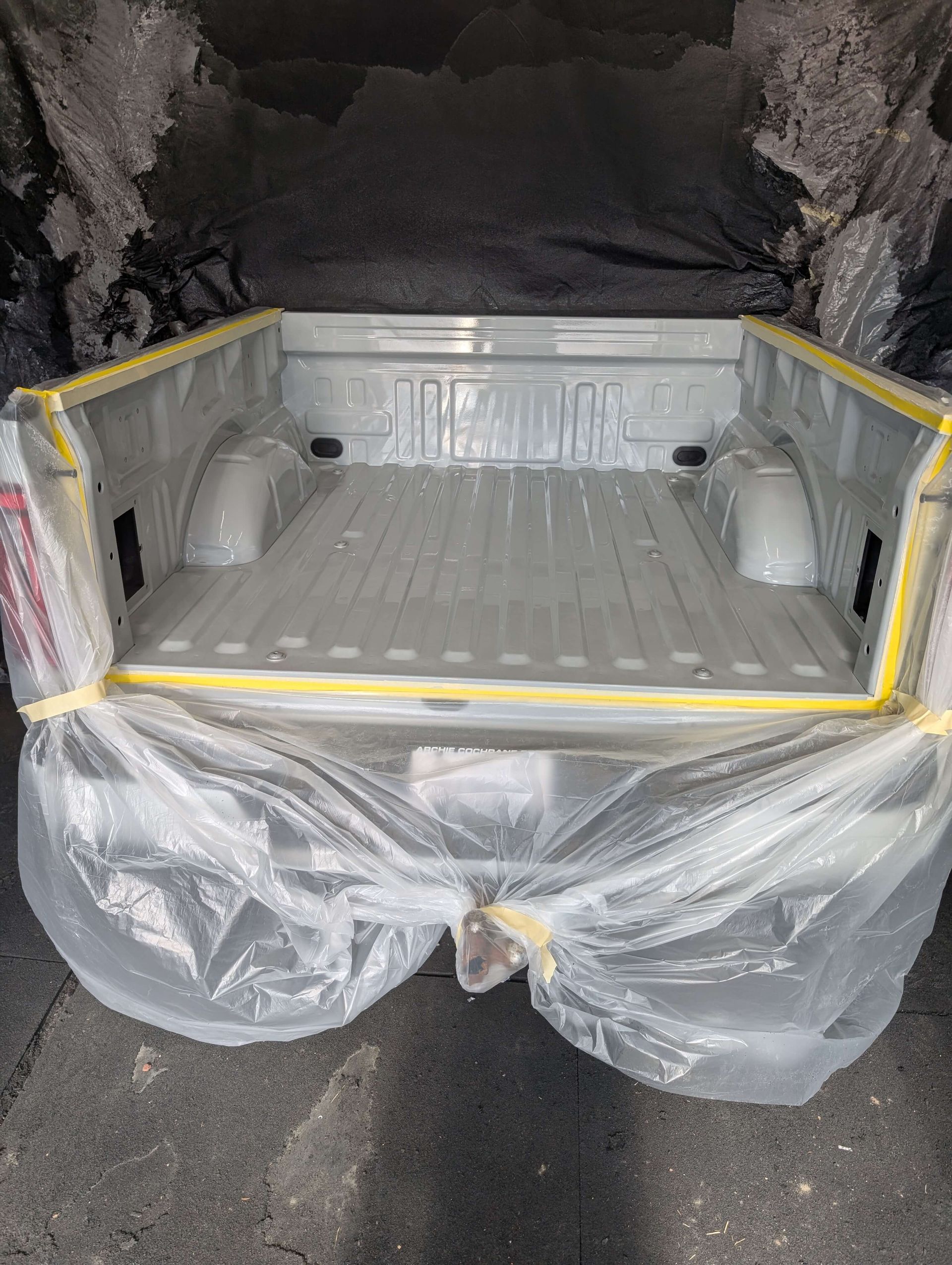 Empty truck bed, primed in gray, taped with yellow tape, covered in plastic.