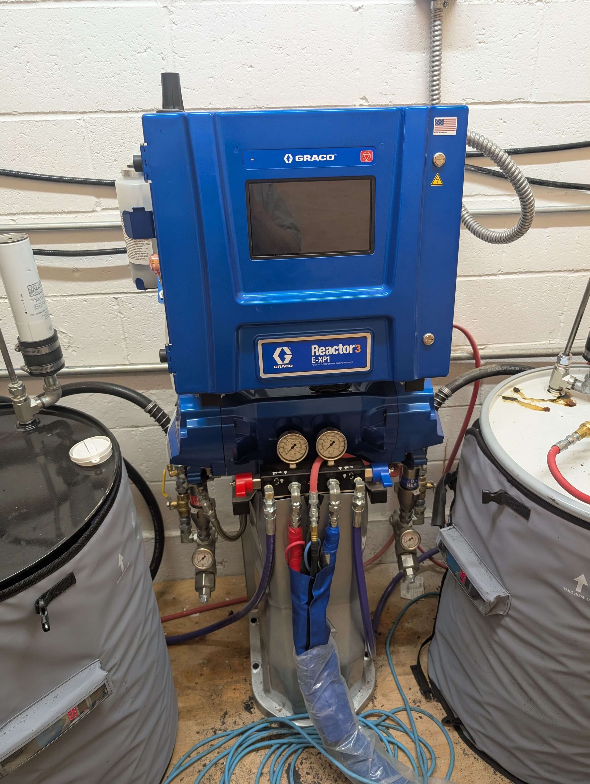 Blue industrial dispensing machine with screen, connected to barrels, in a work setting.