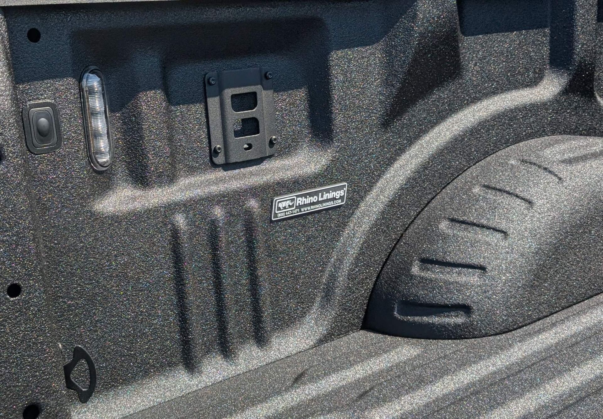 Close-up of a black truck bed with a textured coating and metal fixtures, including a mounting bracket and brand emblem.