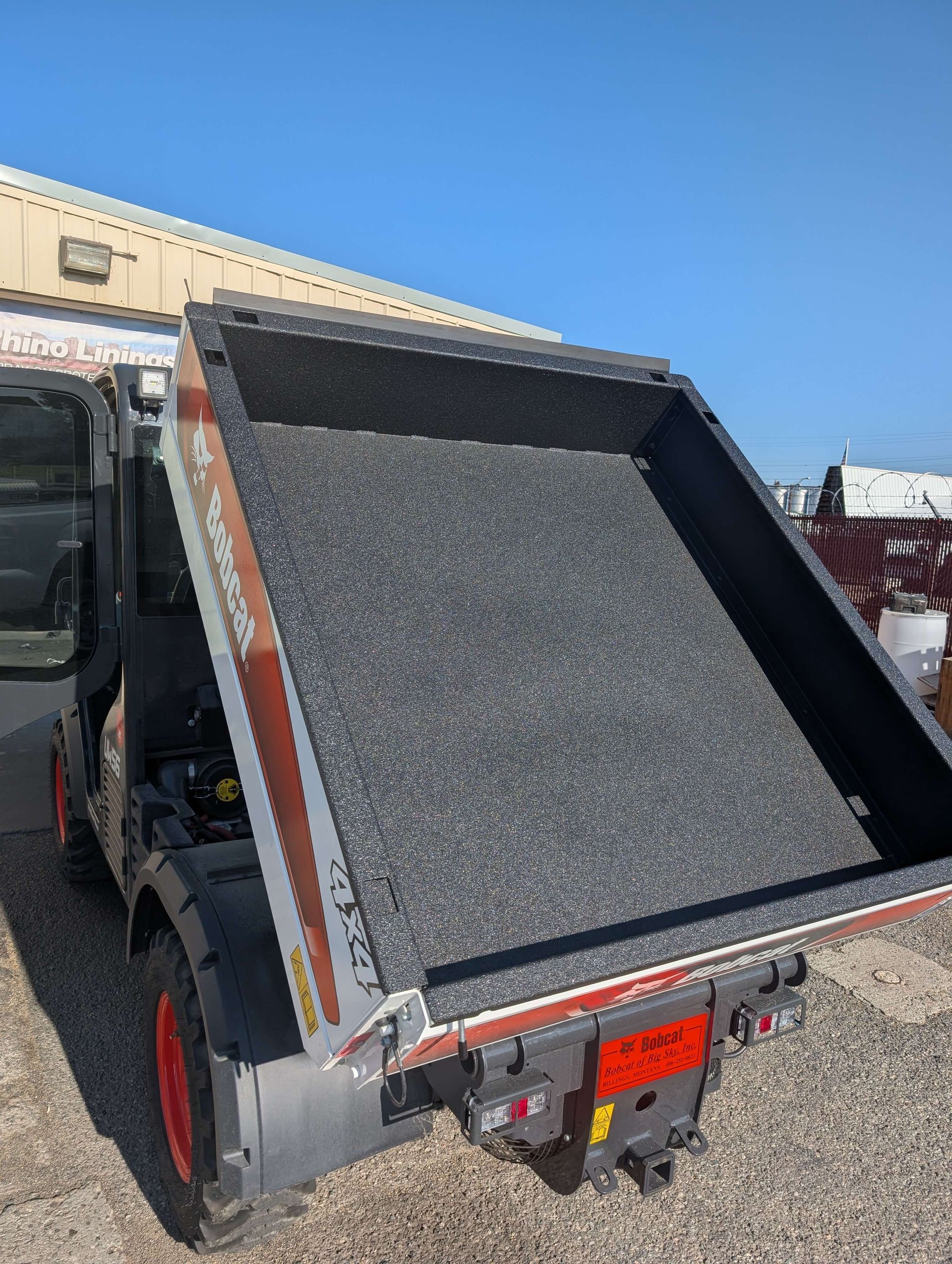 A Bobcat mini-dumper, bed tilted, filled with dark material, outdoors. Sunny day.