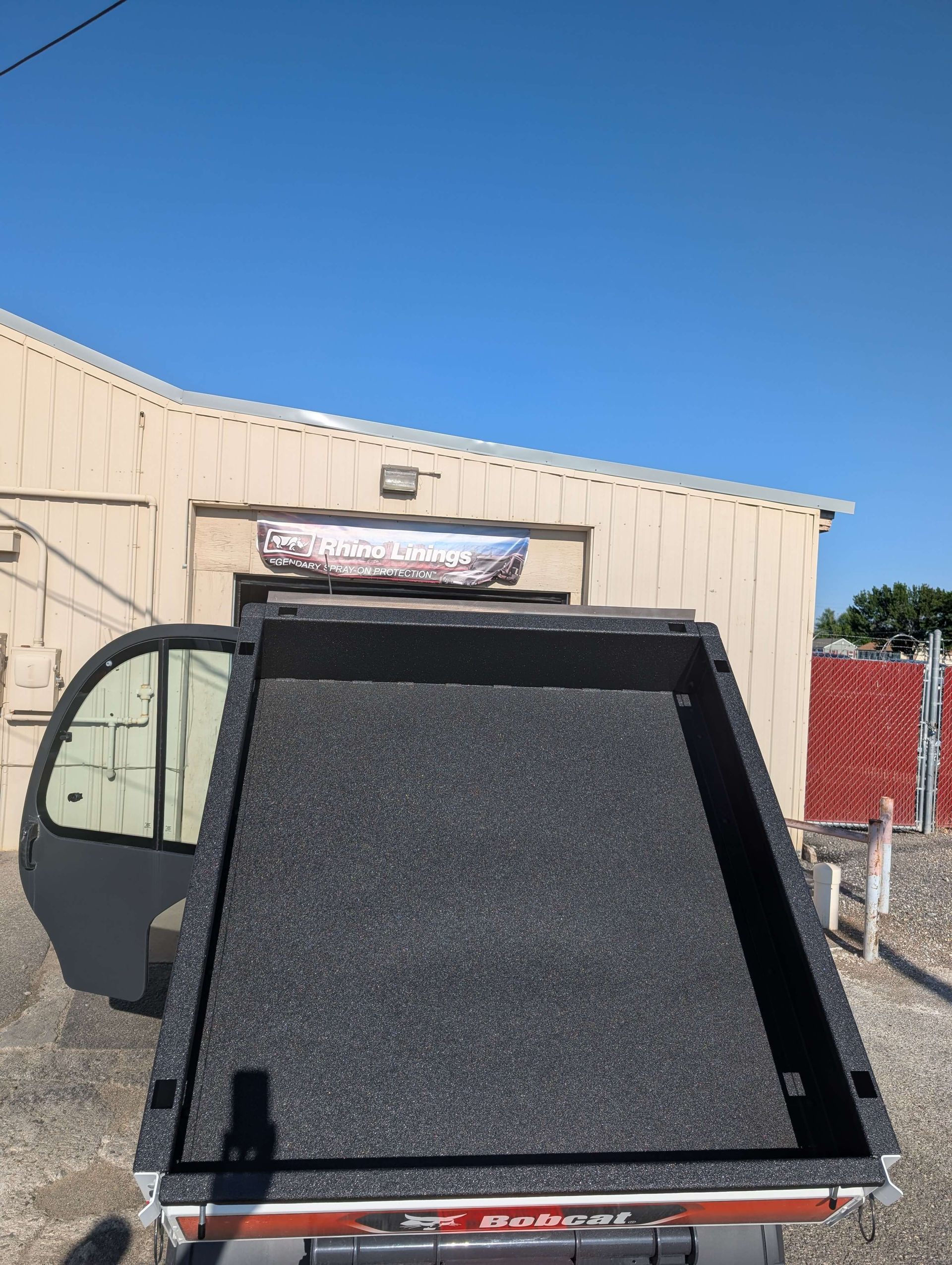 A black truck bed with a textured surface is in front of a building on a sunny day.
