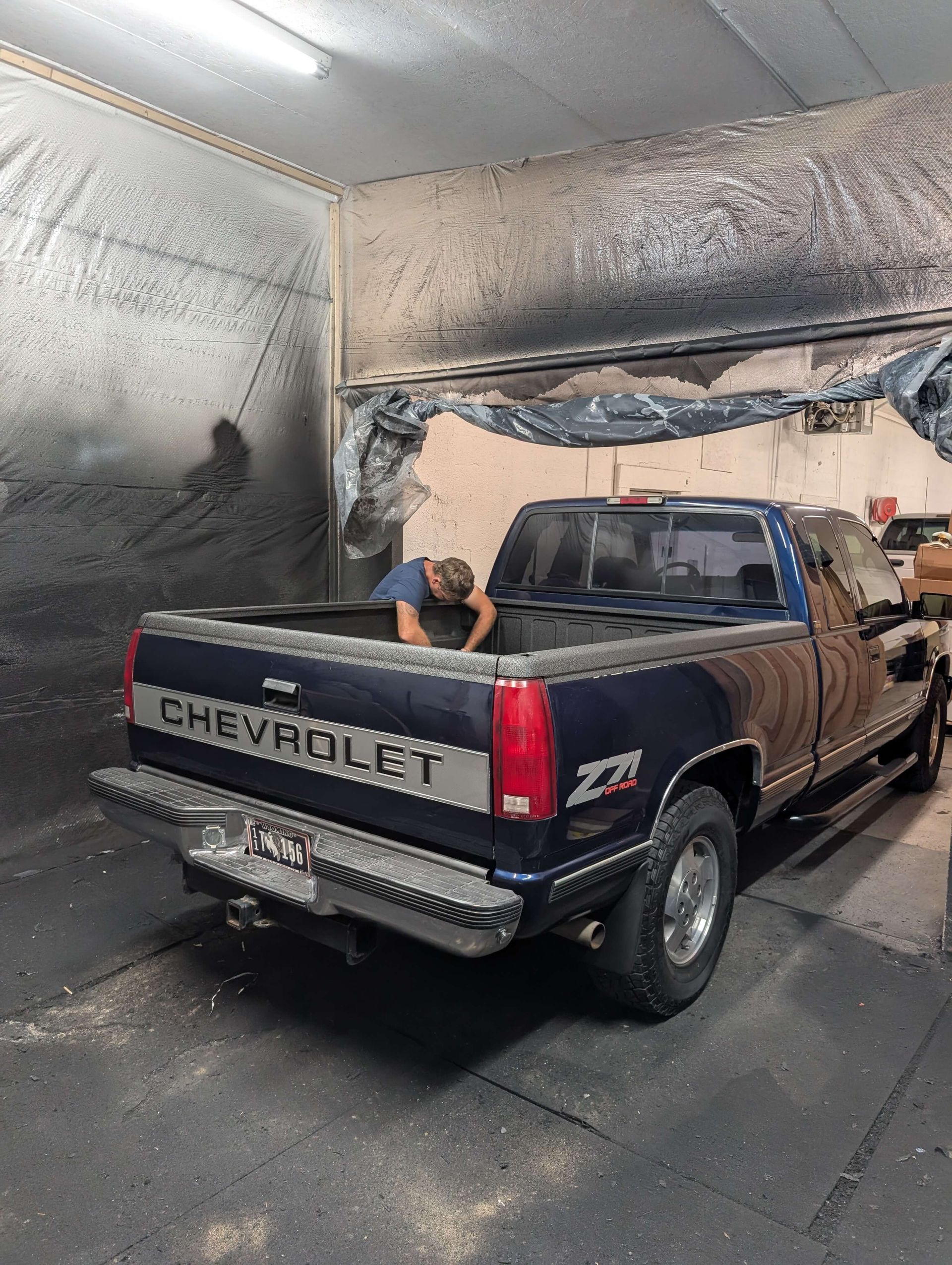 A person leans into a blue Chevy truck bed inside a shop with dark, possibly ashy, walls and ceiling.