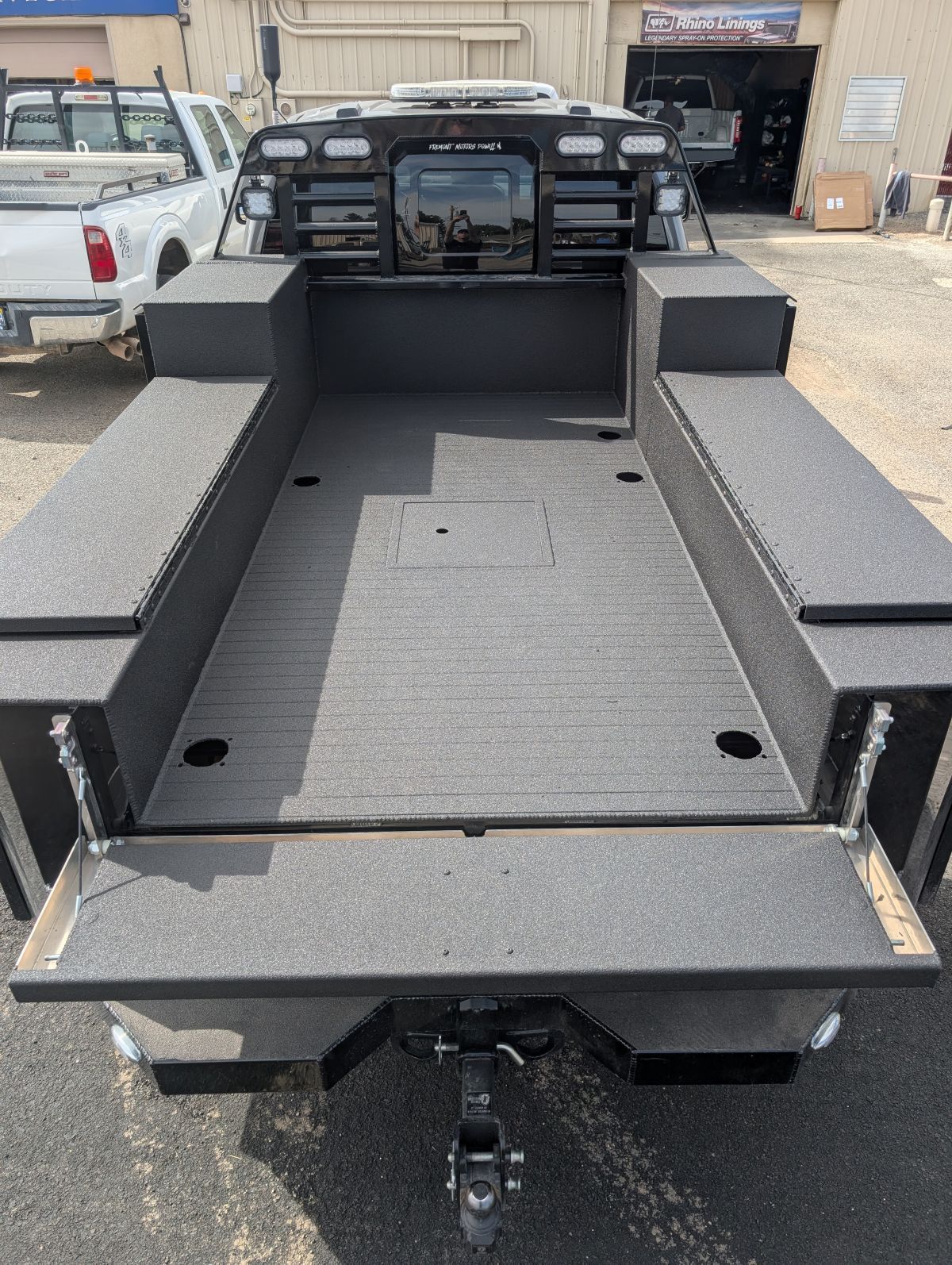 Black flatbed truck bed with side storage compartments and a trailer hitch, outdoors.