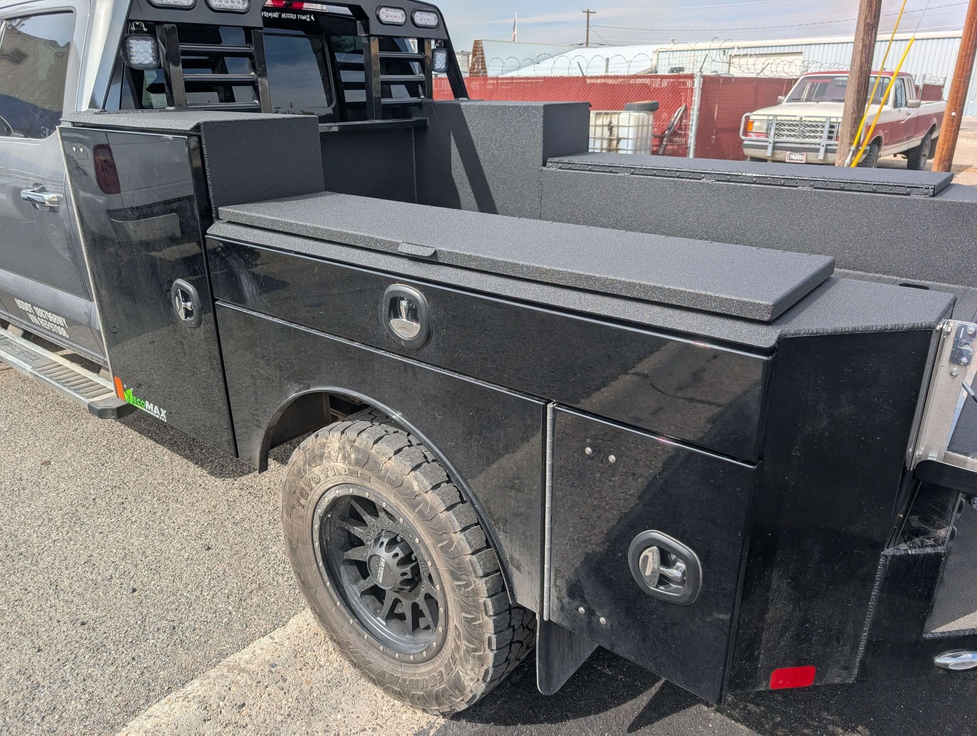 Black truck bed with side toolboxes, a black tire, and a red taillight in an outdoor setting.