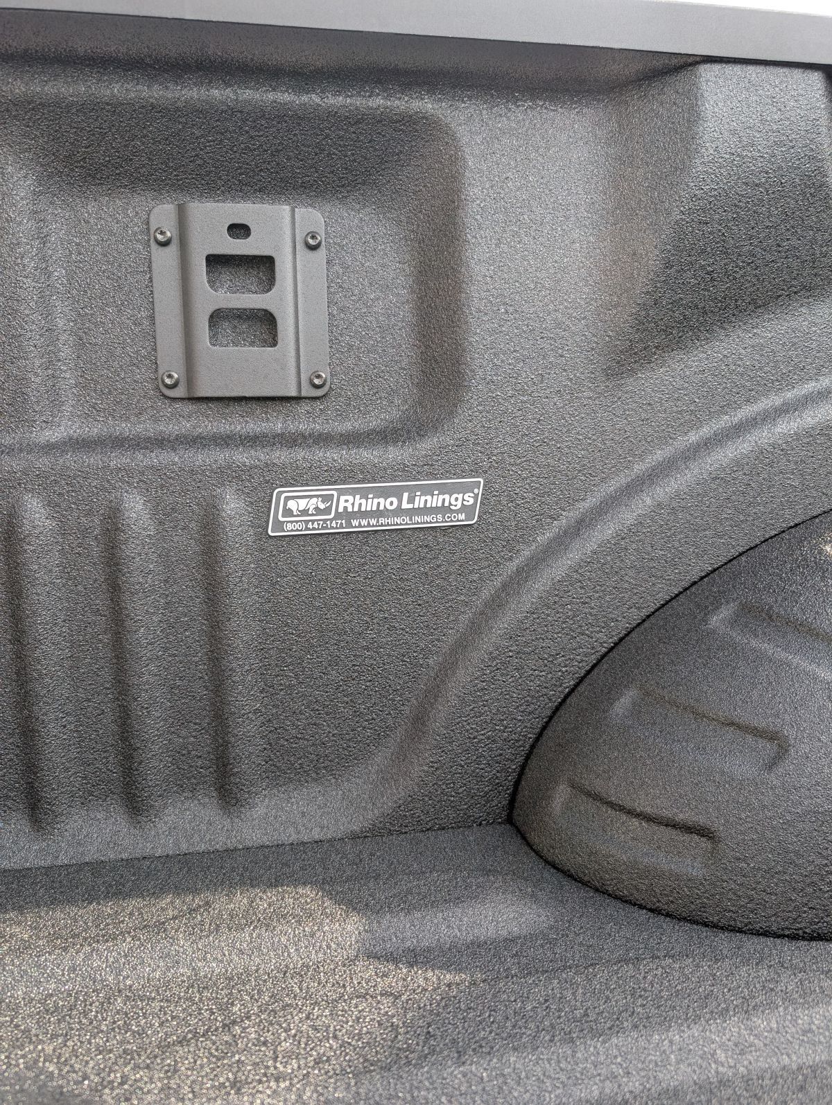 Black truck bed with Rhino Linings spray-on liner.  Mounting bracket and label visible.