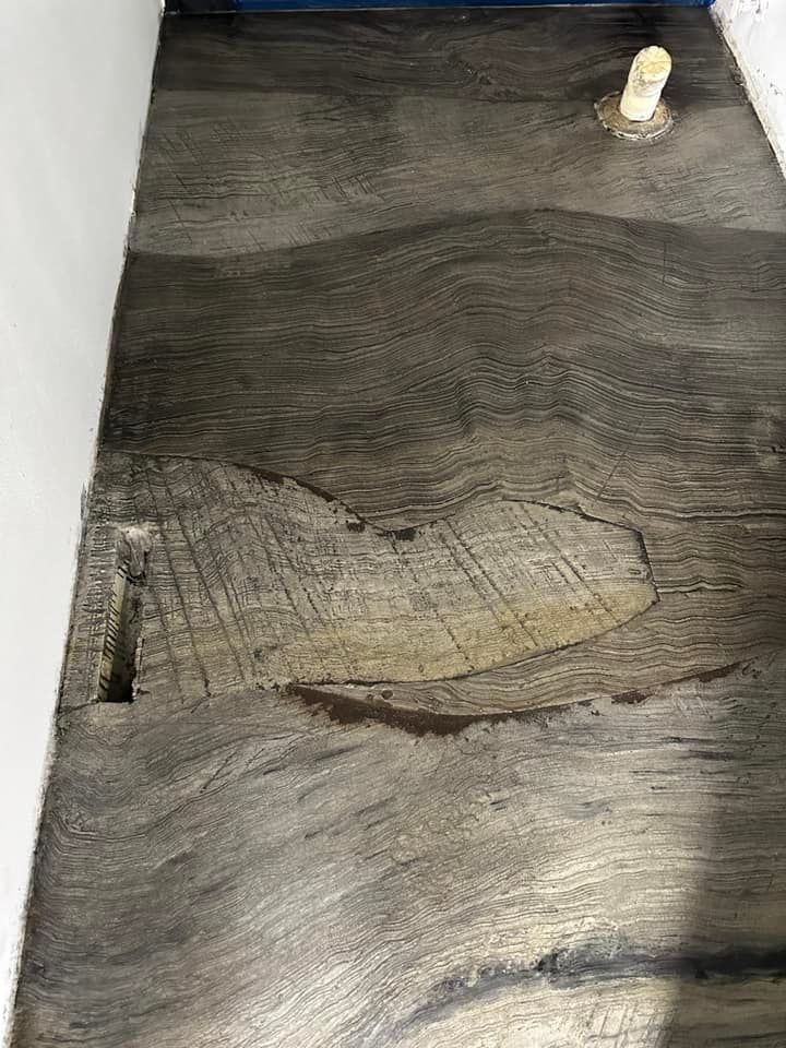 Gray, wood-grain patterned floor with a damaged section and a pipe in the upper right.