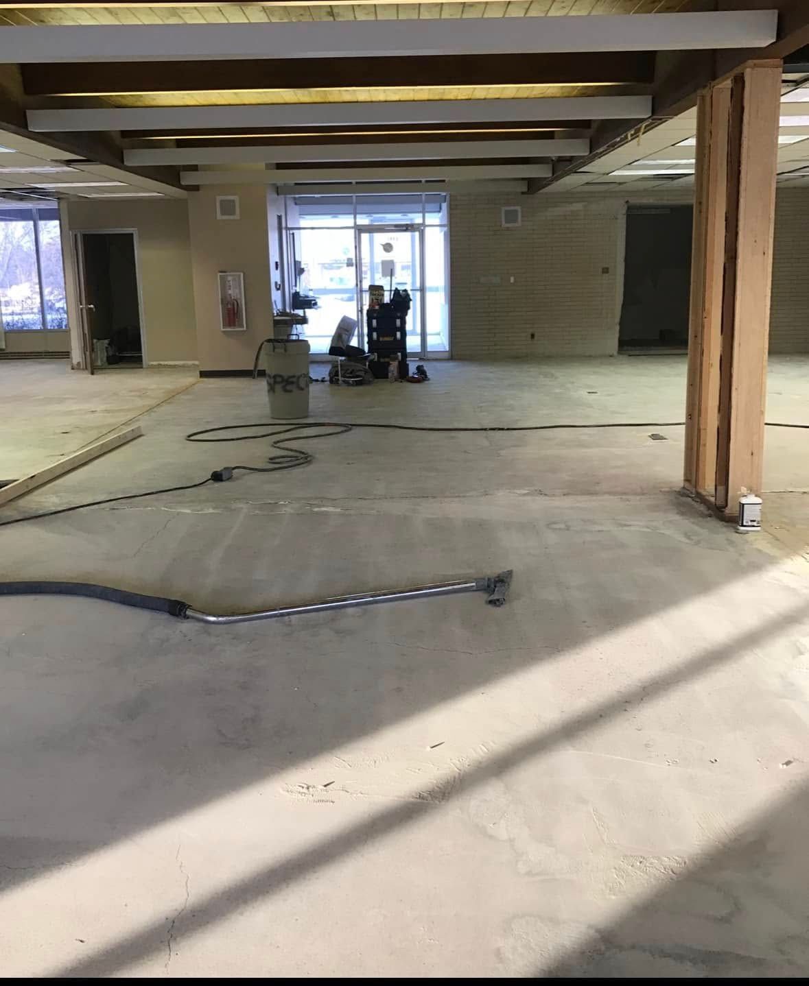 A wide, empty room with exposed beams and concrete floor. Construction equipment by glass doors.
