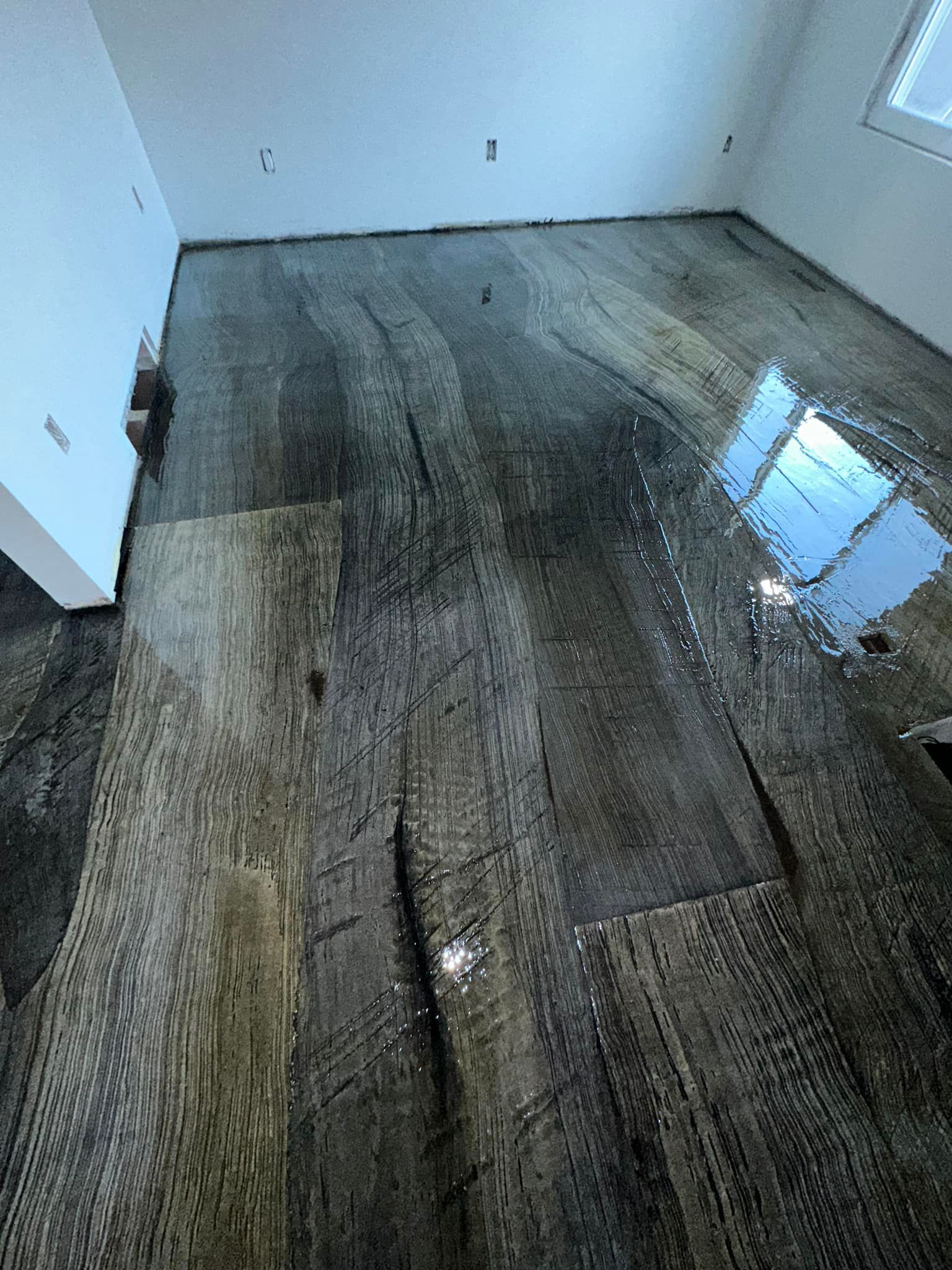 Flooded wood-look flooring in a room with a white wall and a window reflecting the sky.