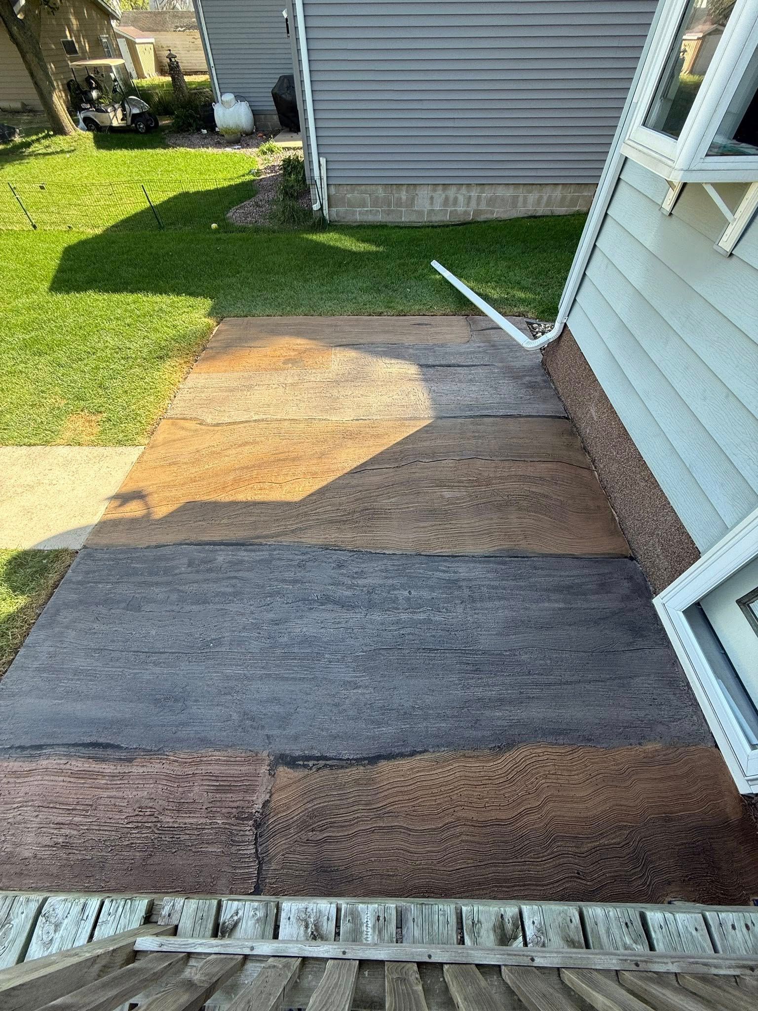 Concrete walkway with textured woodgrain pattern in shades of brown, gray, and tan, next to a house with white trim.