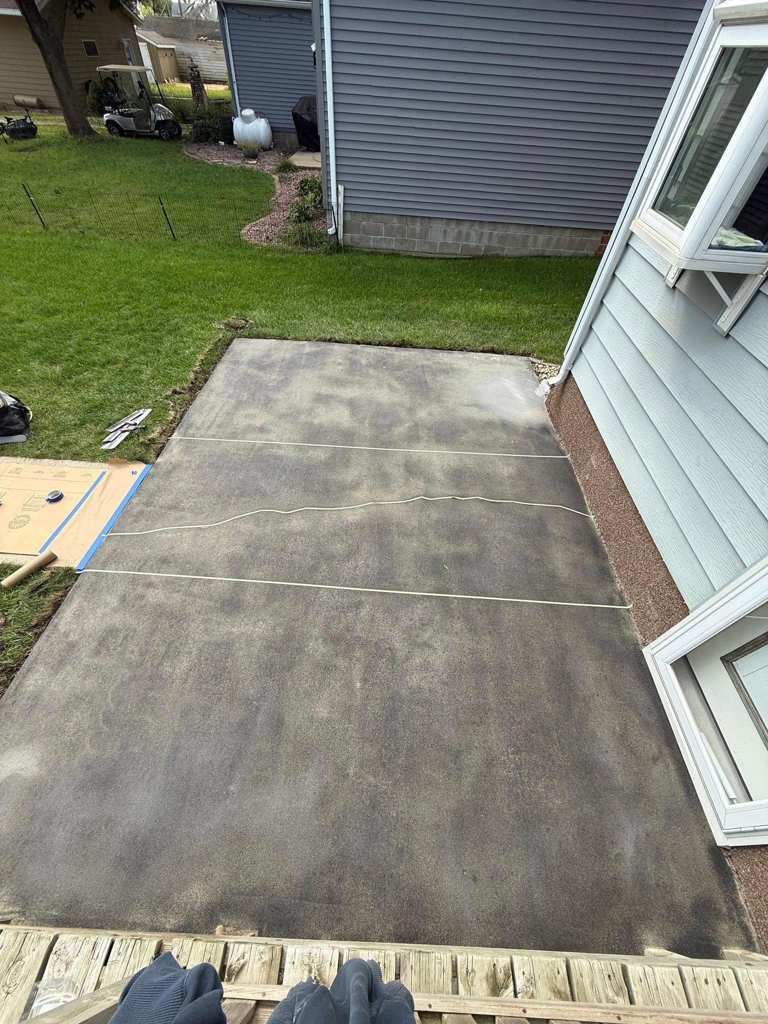 Concrete patio with white lines, next to a house with blue siding and windows. Grass in the background.