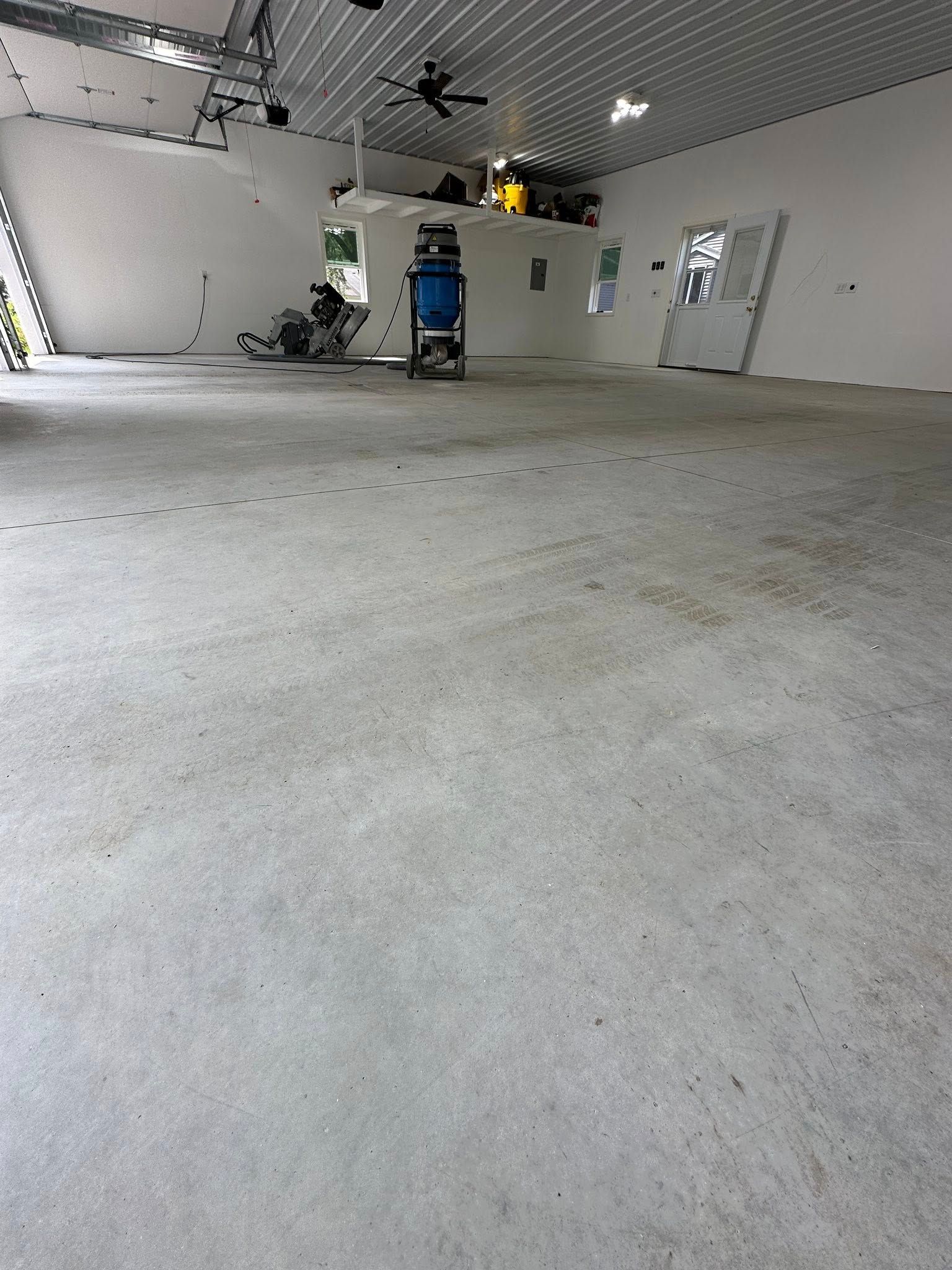 Empty, light-colored concrete floored interior. White walls, high ceiling. Tools and equipment visible in the background.