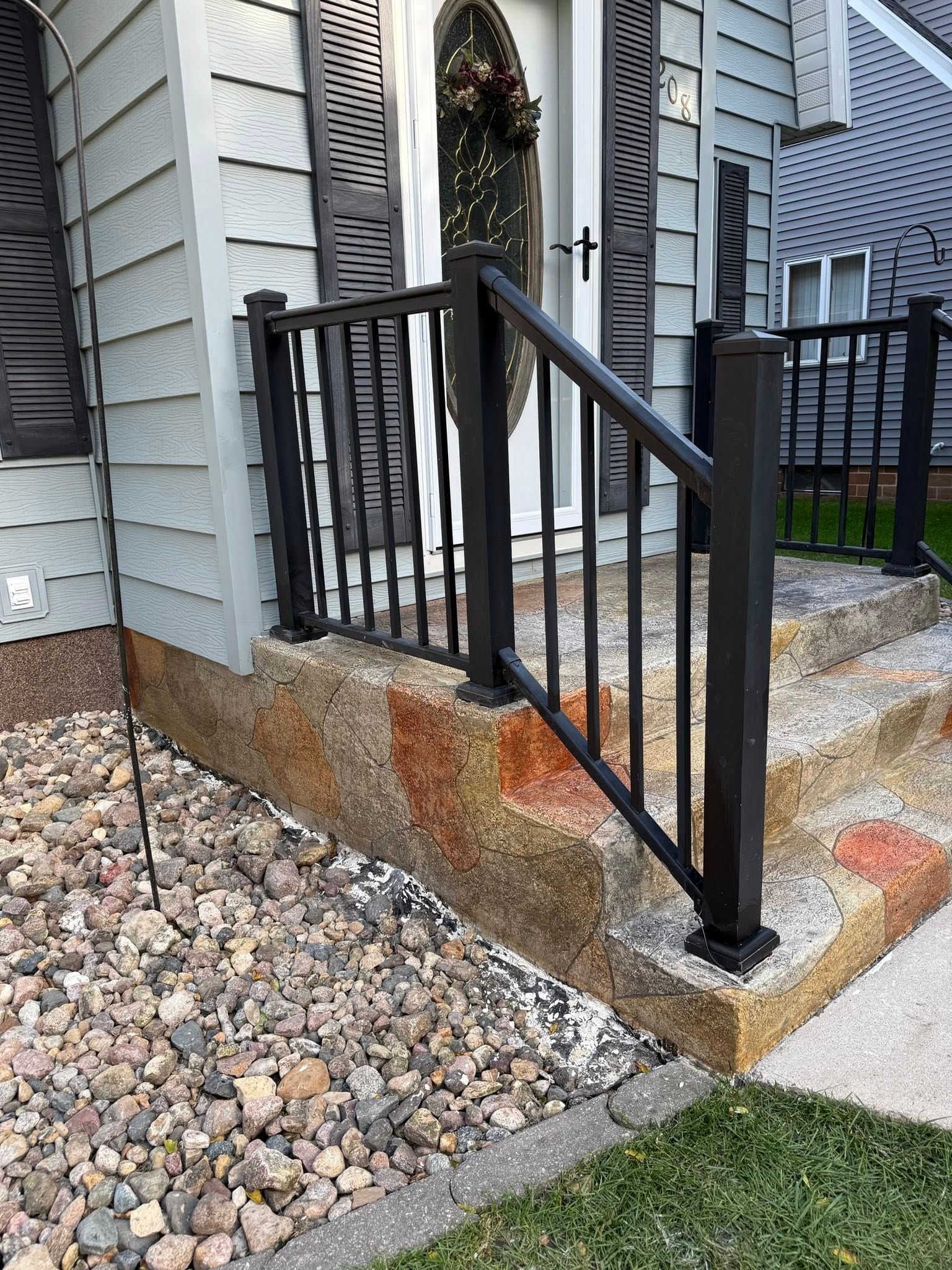 Black railing on a stone-covered porch with steps. Gravel and grass are in the foreground. Gray house siding in the background.
