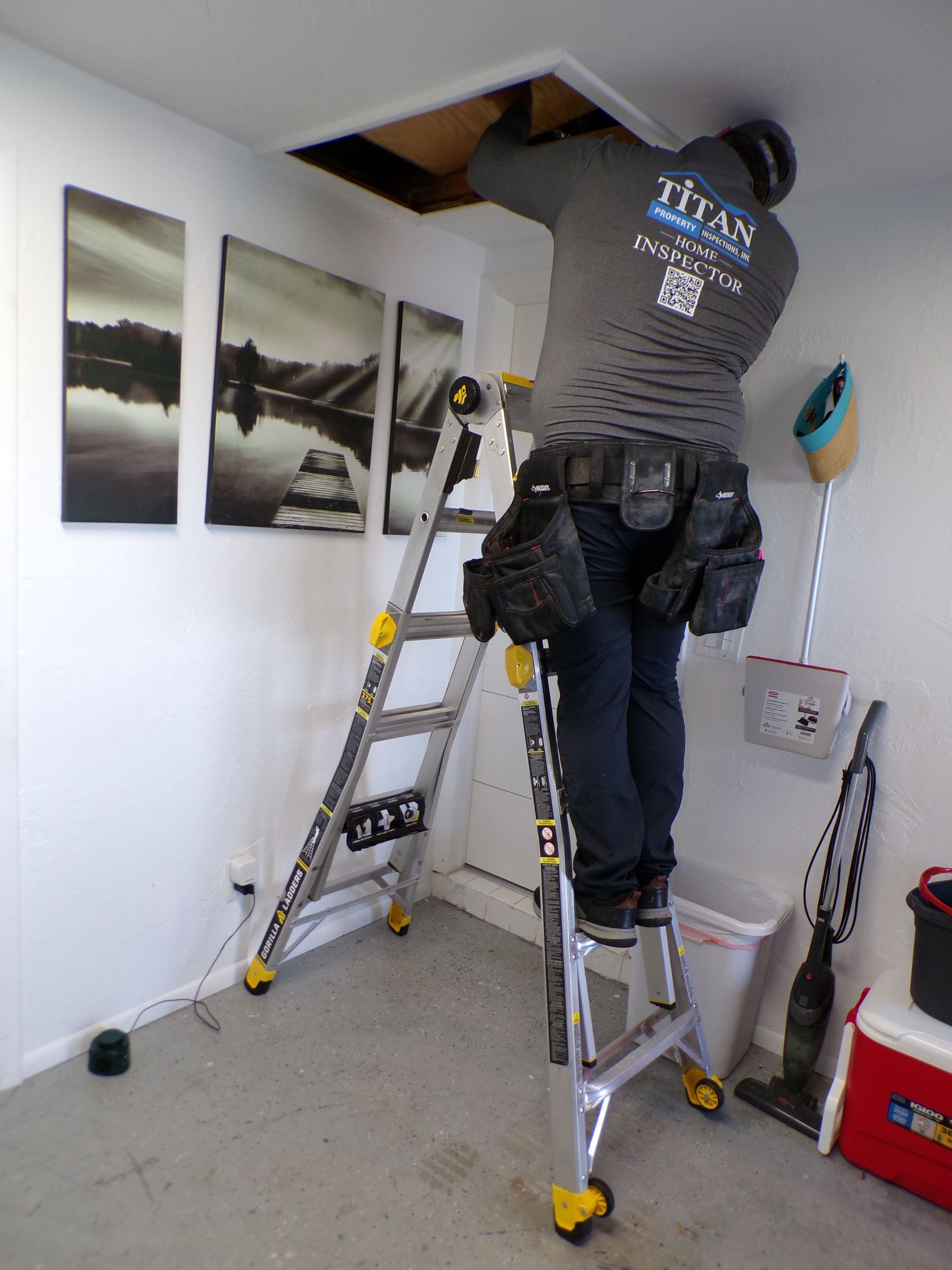 Persona en escalera trabajando en el acceso al techo, con cinturón de herramientas. Habitación con arte, aspiradora y cubo de basura.