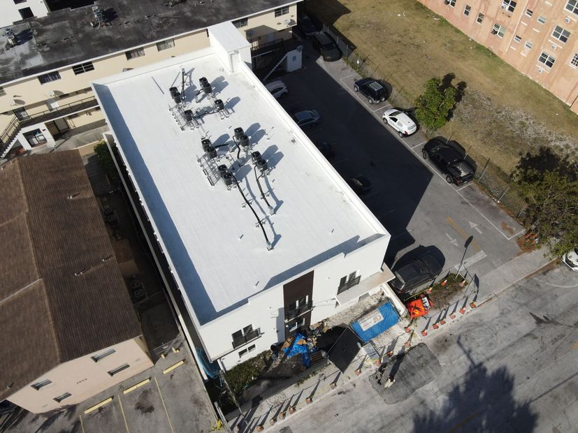 Aerial view of a white rectangular building with rooftop equipment; cars parked nearby on a paved lot and street.