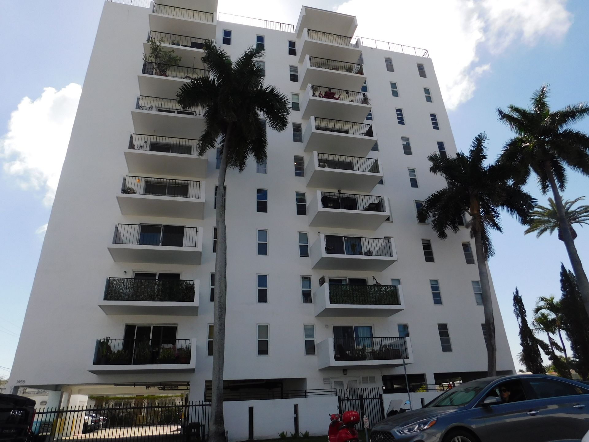 Edificio de apartamentos blanco de varios pisos con balcones, palmeras y autos estacionados.