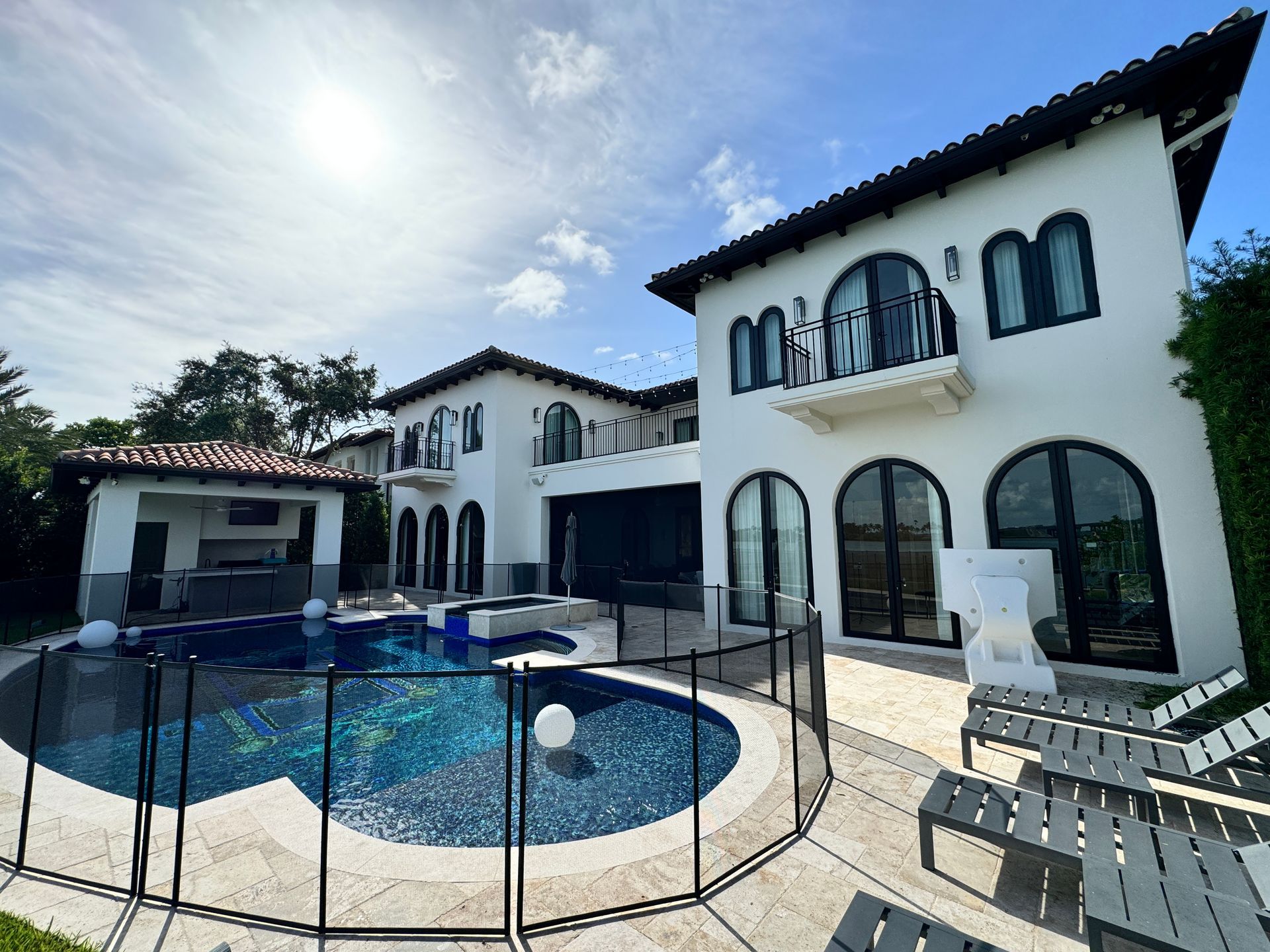 Lujosa mansión blanca con piscina, cocina al aire libre y muebles de patio bajo un cielo brillante.