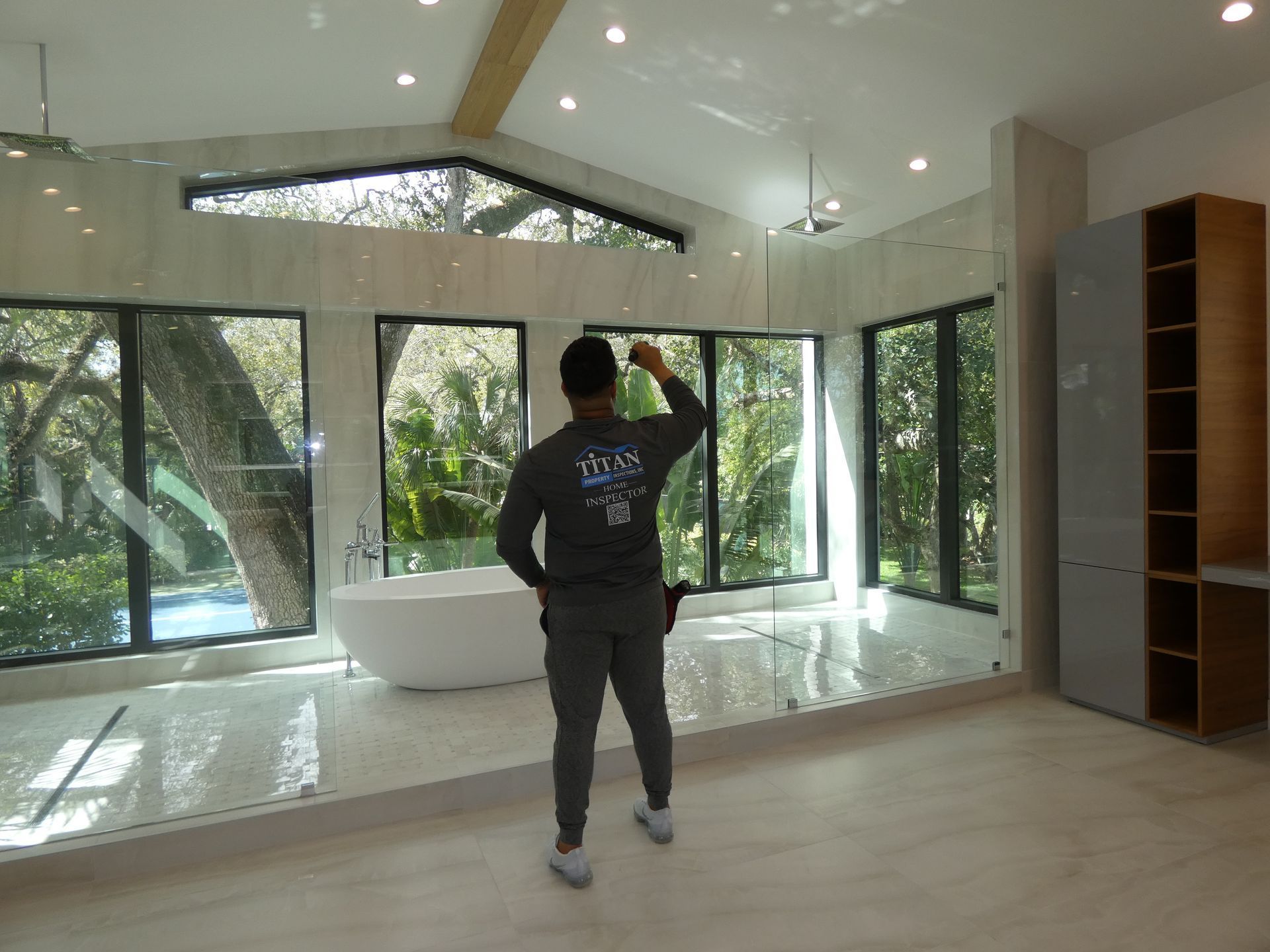 Un hombre vestido de gris inspecciona un baño moderno con ventanas grandes, una bañera independiente y acabados de colores neutros.