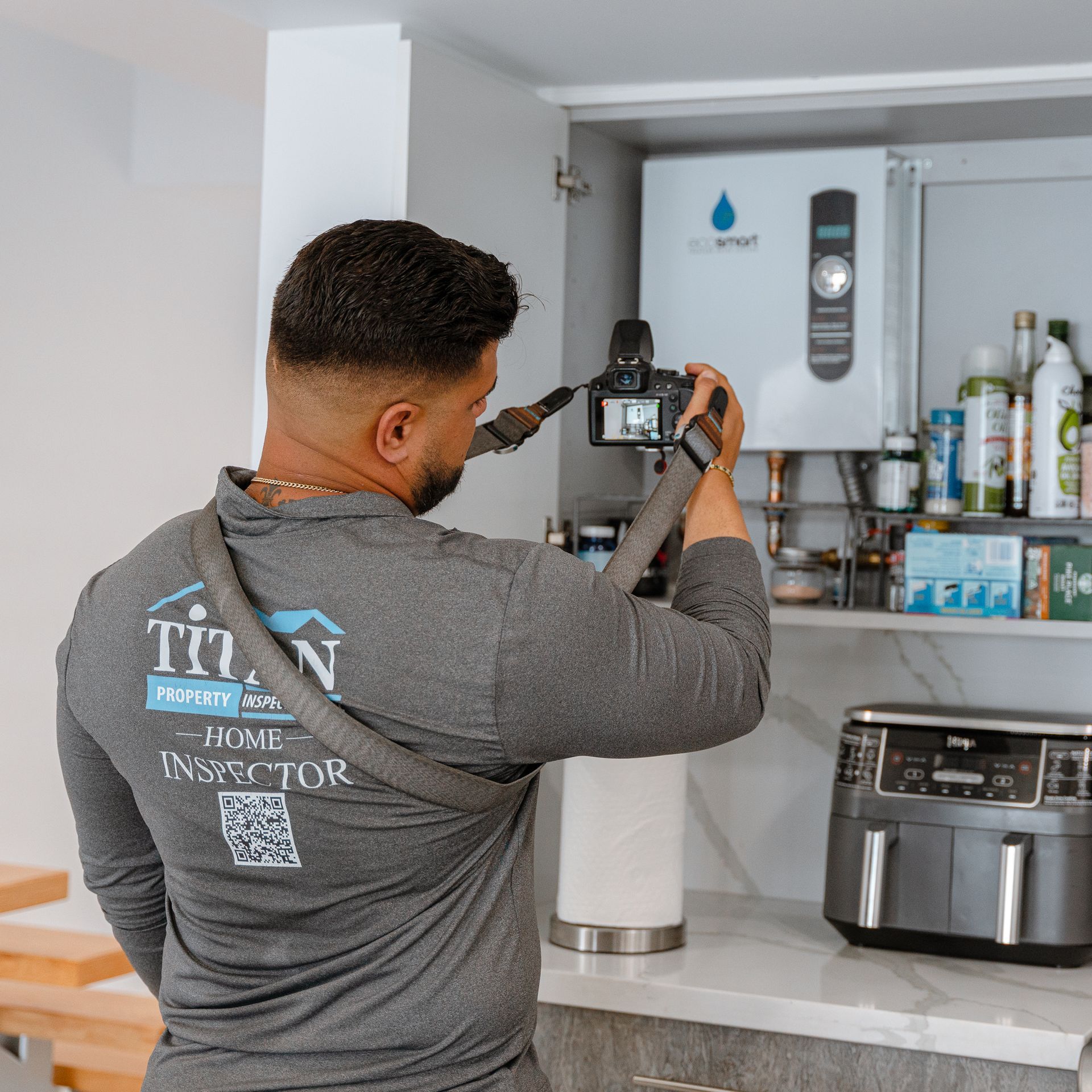 Un inspector de viviendas toma una fotografía de un calentador de agua dentro de un gabinete de cocina, vistiendo una camisa gris, al lado de una freidora de aire.