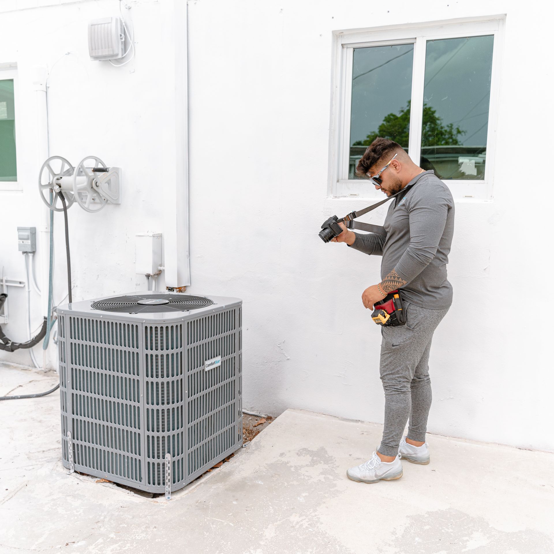 Hombre con mono gris fotografiando una unidad de aire acondicionado exterior cerca de un edificio blanco.