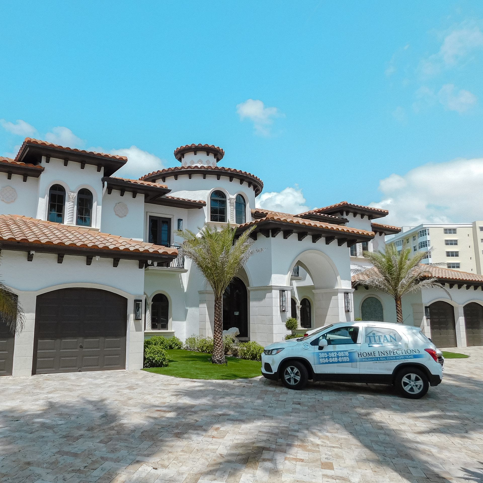 Un coche blanco aparcado delante de una gran casa blanca con tejas marrones bajo un cielo azul.