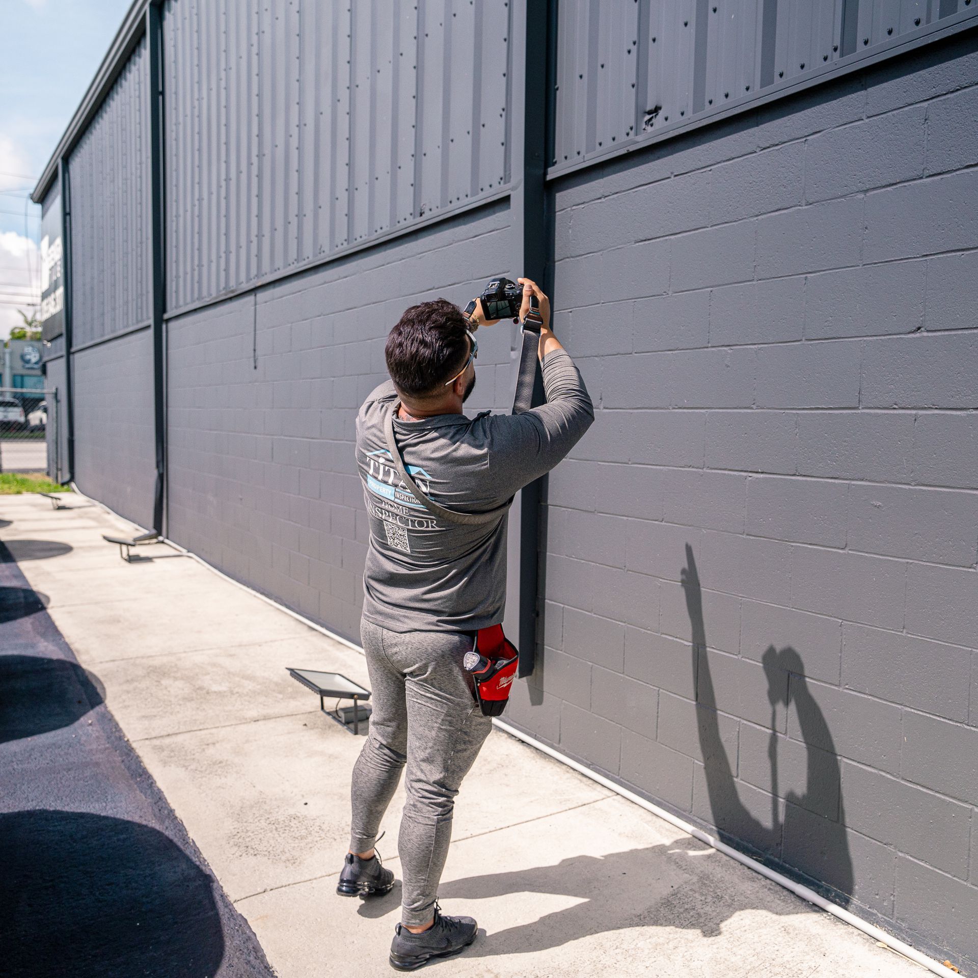 Hombre con ropa deportiva gris fotografiando una pared gris oscura. Sombra proyectada en la pared. Luz solar intensa.