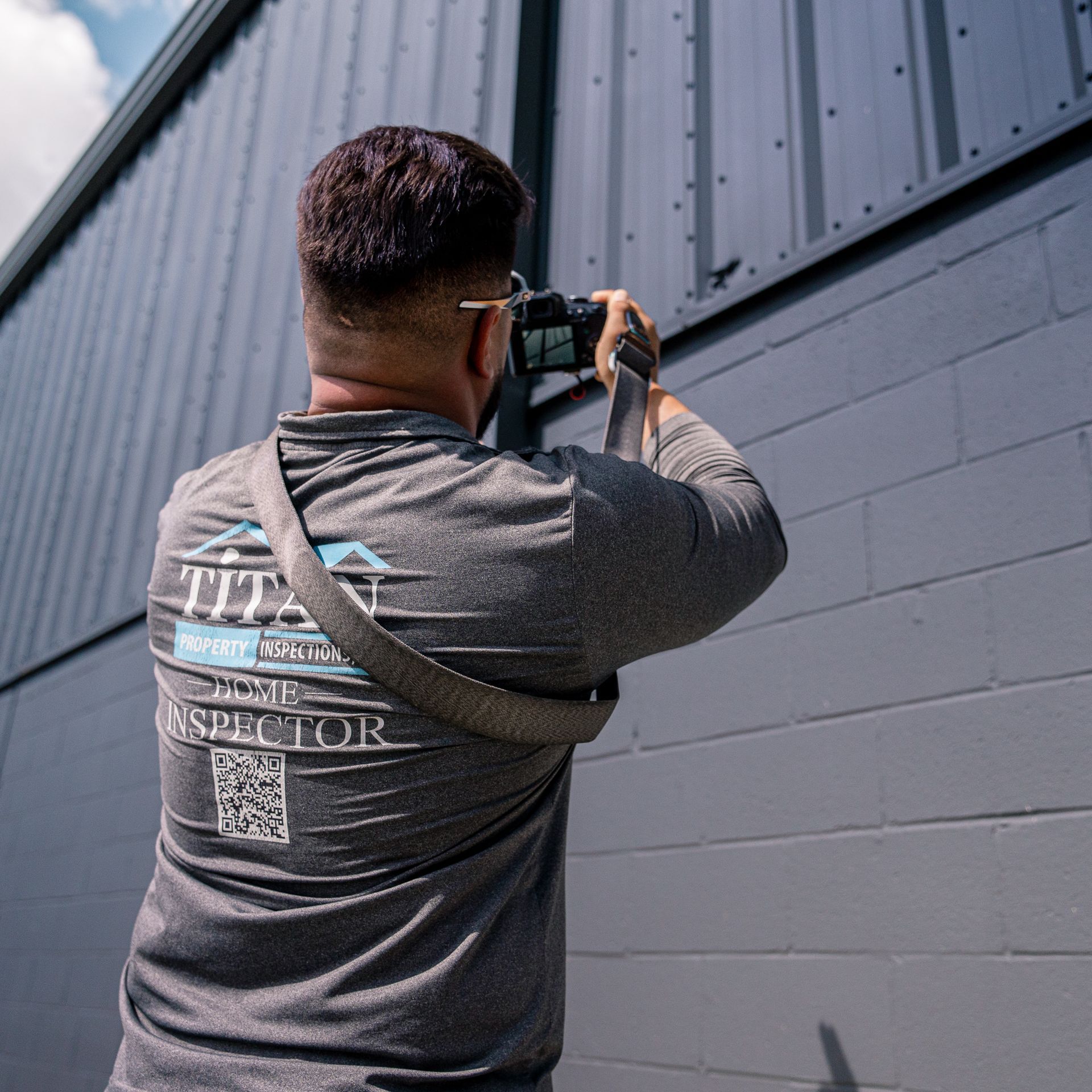 Persona tomando una foto de un edificio gris. Lleva una camiseta con la inscripción