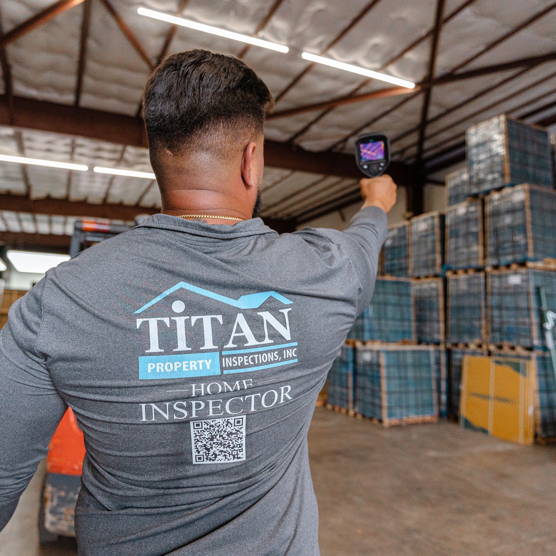 Persona con camisa gris de Titan Property utiliza un dispositivo portátil en un almacén para inspeccionar pallets apilados.