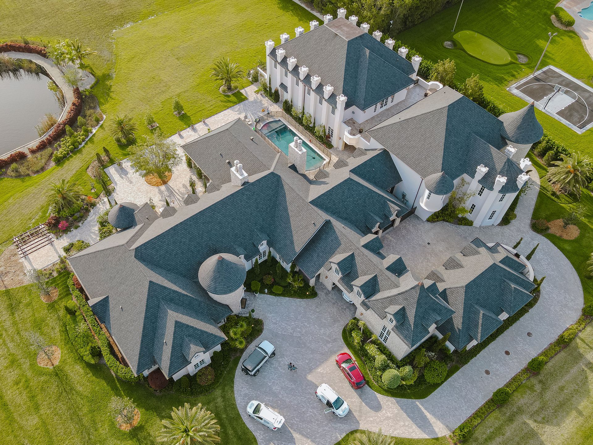 Vista aérea de una casa grande con un tejado complejo, piscina y entrada circular. Varios vehículos estacionados.