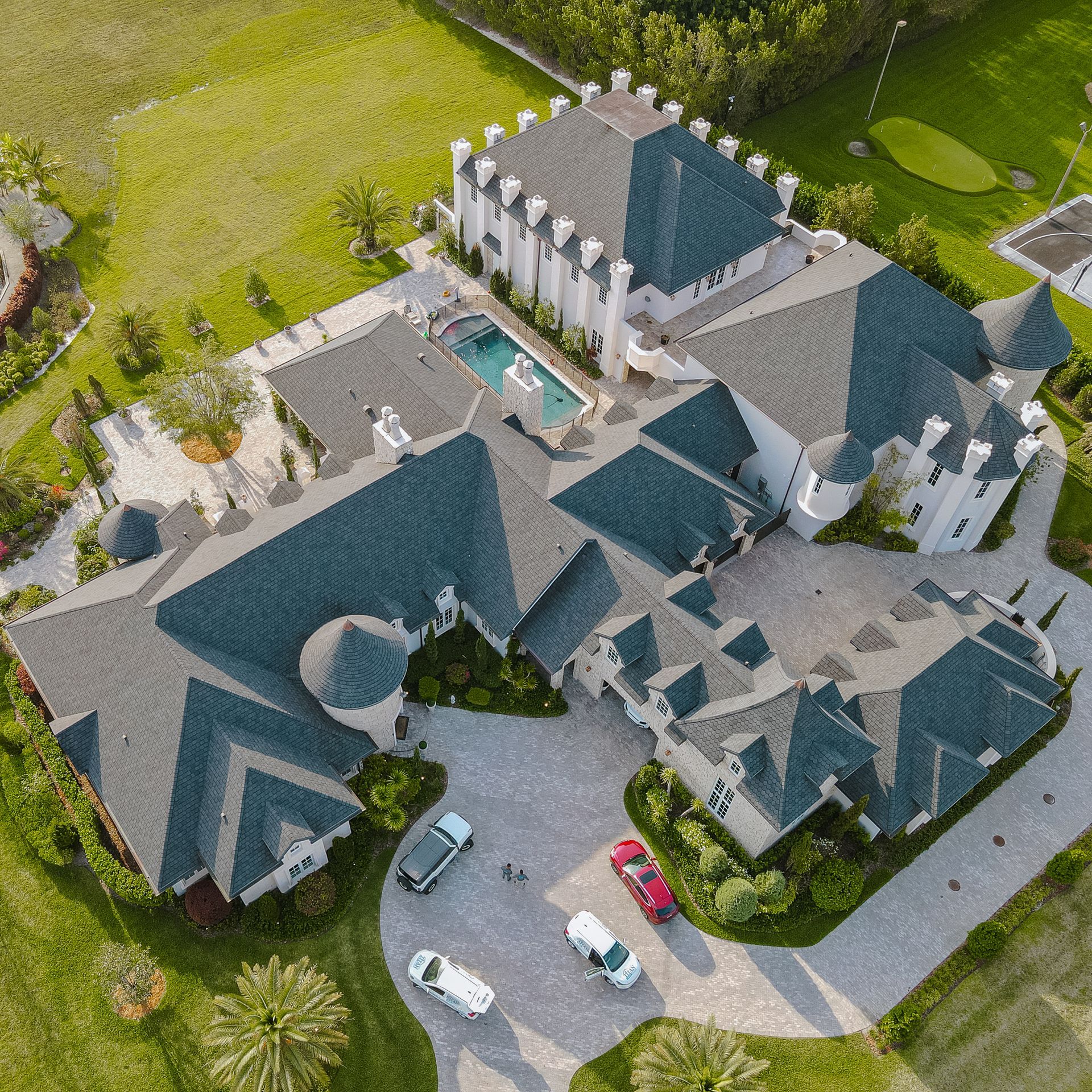 Vista aérea de una casa grande con un techo complejo y una entrada circular; los autos están estacionados afuera, cerca de una piscina.